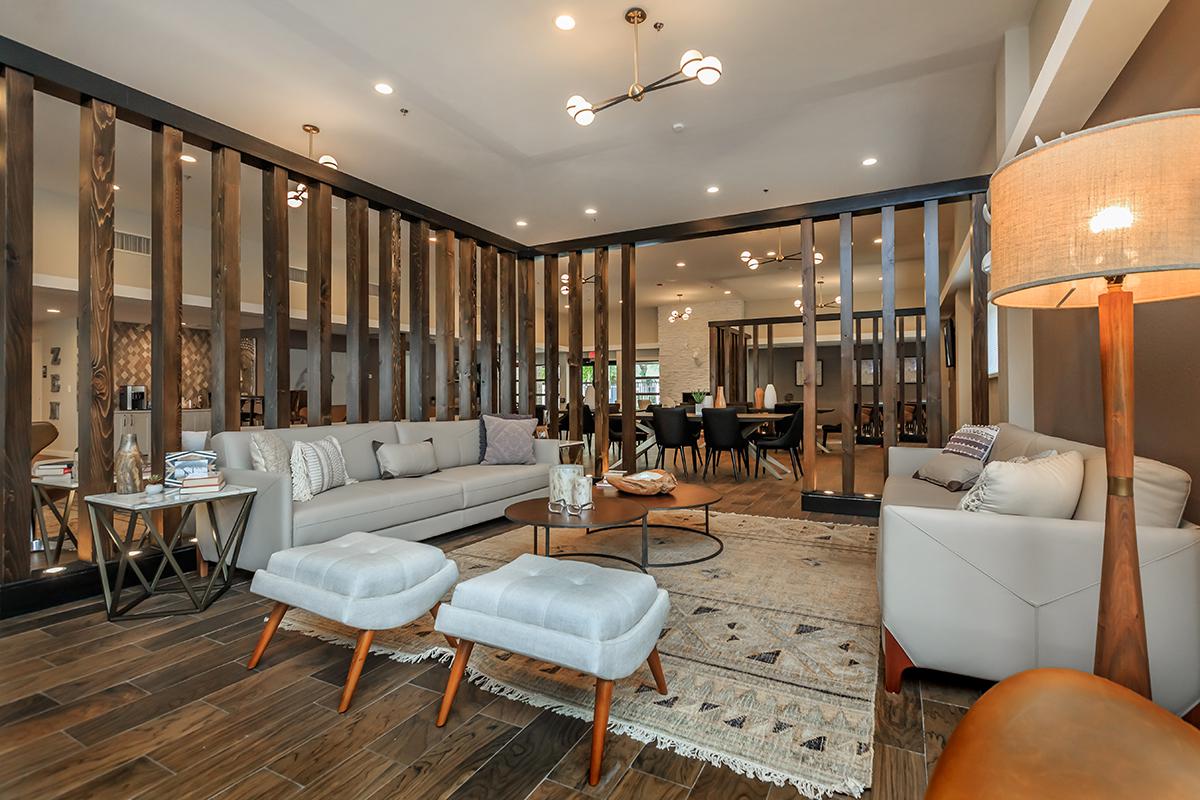 A modern and spacious living area featuring a light-colored sofa, two ottomans, a round coffee table, and an area rug. The room is divided by wooden partitions and has stylish lighting fixtures. In the background, there are dining tables and chairs, creating an inviting atmosphere.