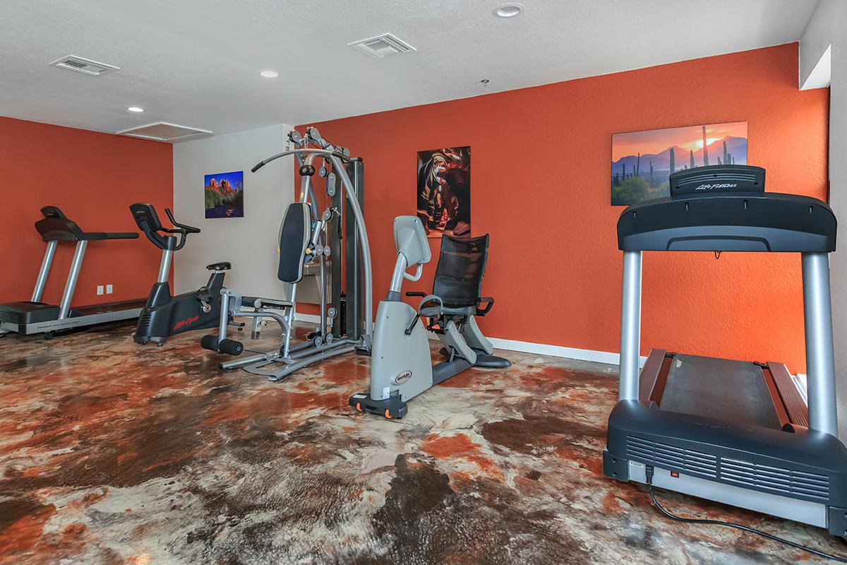 A gym area featuring exercise equipment including a treadmill, strength training machines, and stationary bikes, with orange walls and artwork in the background.