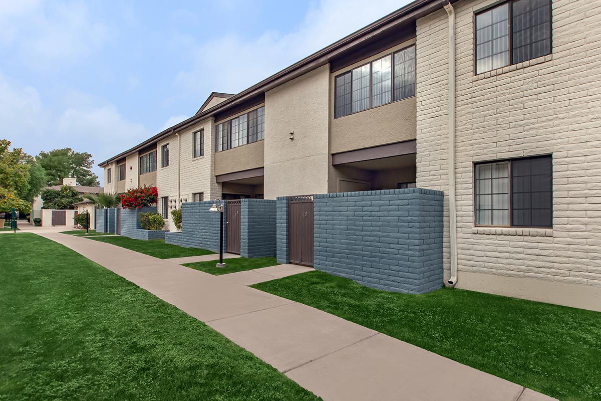 A view of a multi-unit residential building with light-colored walls, featuring several windows. The building is bordered by neatly trimmed grass and a wide concrete pathway. In the foreground, there are blue-painted walls and doors, along with landscaped flowerbeds adorned with red flowers.