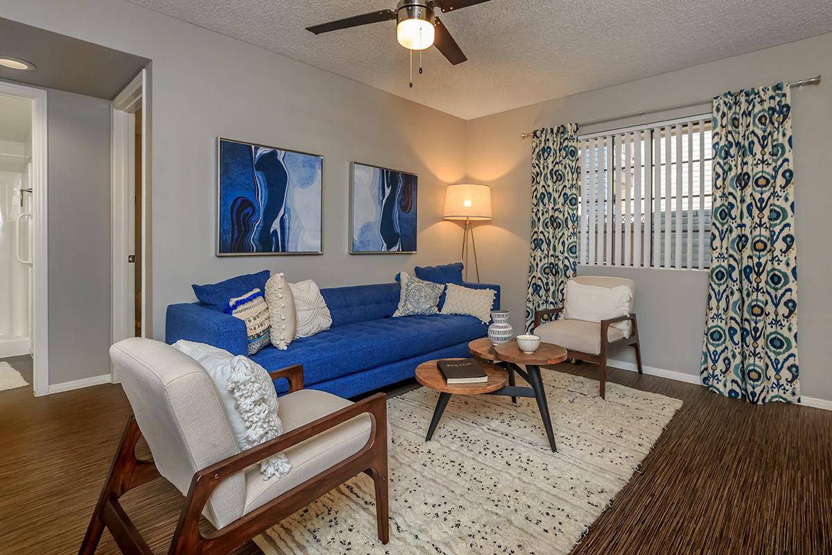 A cozy living room featuring a blue sofa adorned with decorative pillows, two stylish armchairs, and a round coffee table. The room has a light-colored area rug, modern artwork on the walls, and a ceiling fan. Bright natural light filters through a window with patterned curtains.