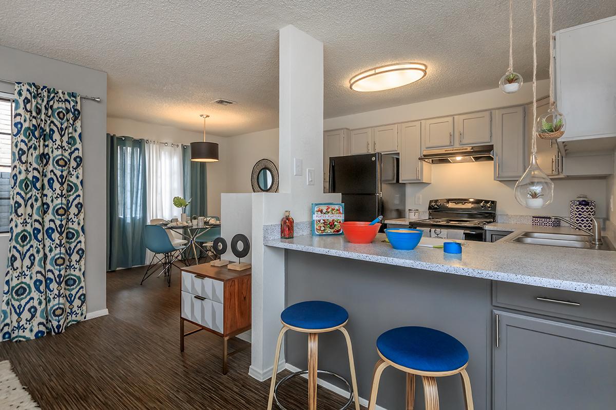 A modern kitchen with a gray and white color scheme featuring a breakfast bar with two blue stools, a stainless steel stove, and a refrigerator. The dining area in the background has a round table with blue chairs and decorative curtains. Bright natural light filters in through the window.