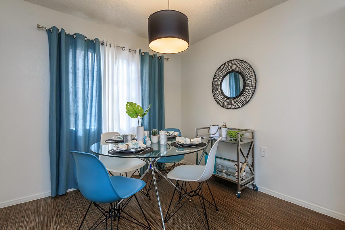 A modern dining area featuring a glass table set for four with blue and white dishware. Two blue chairs are positioned around the table. A large window with light curtains lets in natural light, complemented by a pendant light overhead. A round mirror and a cart are in the corner, adding to the decor.