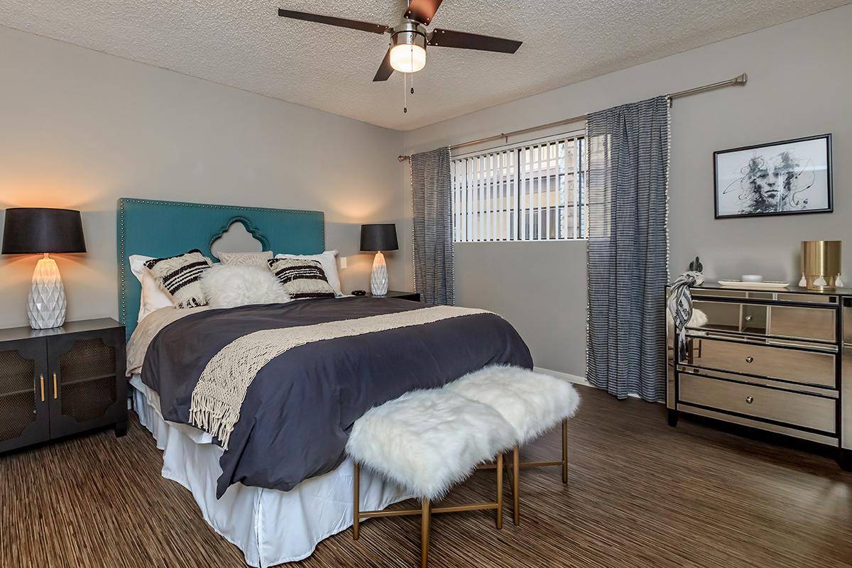 A cozy bedroom featuring a large bed with a dark gray blanket and decorative pillows. There are two stylish lamps on nightstands, a contemporary dresser with metallic accents, and a plush, fur-covered bench at the foot of the bed. Large windows with vertical blinds let in natural light, enhancing the room's warmth.