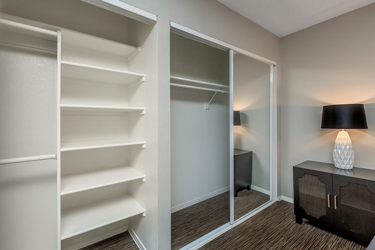 A well-organized closet featuring multiple white shelves and mirrored sliding doors. A stylish lamp stands next to a dark cabinet on the floor, providing a modern and functional space. The walls are painted a soft color, and the flooring is a textured, neutral tone.