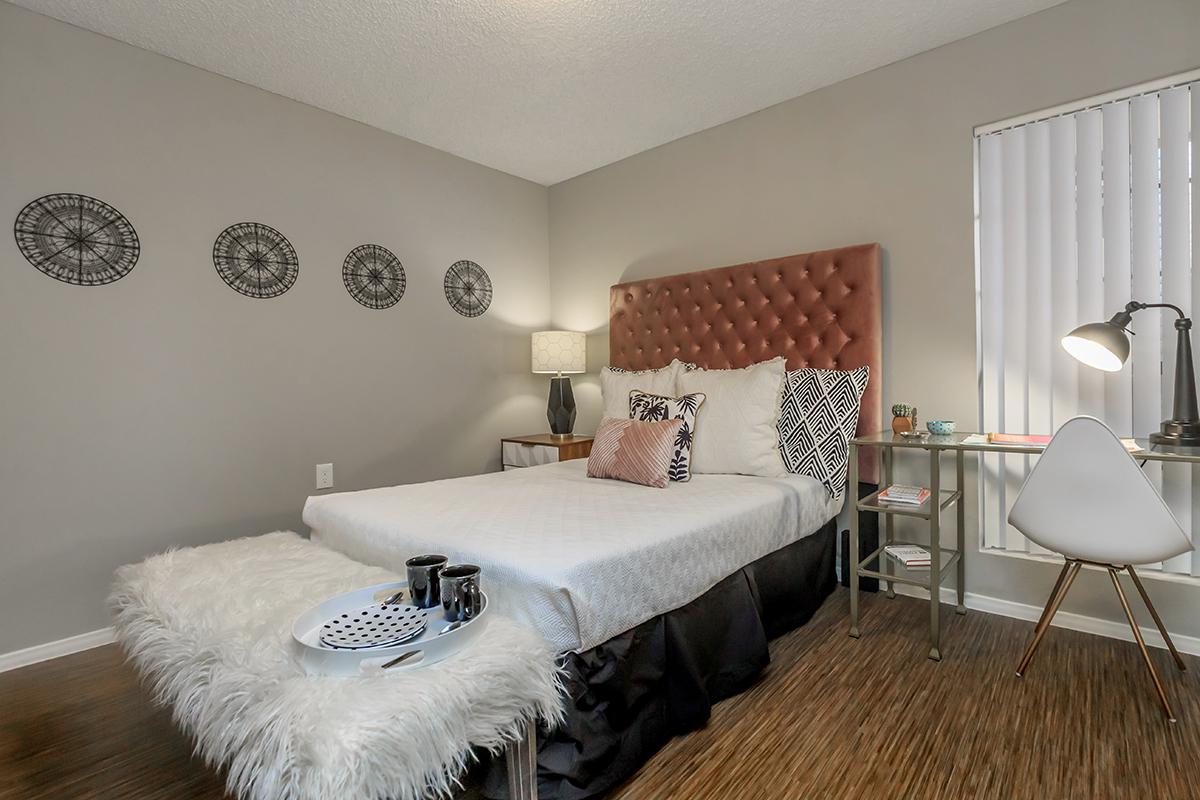 Cozy bedroom featuring a queen-sized bed with a tufted headboard, a stylish bedside lamp, and decorative wall art. A small desk with a lamp and a chair is positioned near a window. A white fur throw sits at the foot of the bed, adding a touch of warmth to the modern decor.