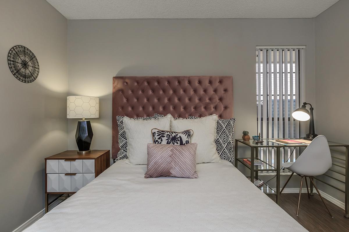 A cozy bedroom featuring a queen-sized bed with a tufted pink headboard, adorned with decorative pillows. There's a stylish nightstand with a black lamp beside it, and a desk with a lamp and colorful stationery. Vertical blinds let in natural light, and a wall clock adds a modern touch.