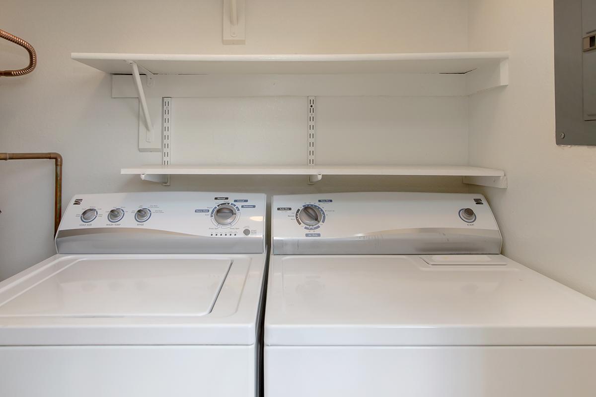 A laundry room featuring a washing machine and dryer side by side. Above them is a white shelf mounted on the wall, providing storage space. The walls are painted a light color, enhancing the clean and organized look of the space.