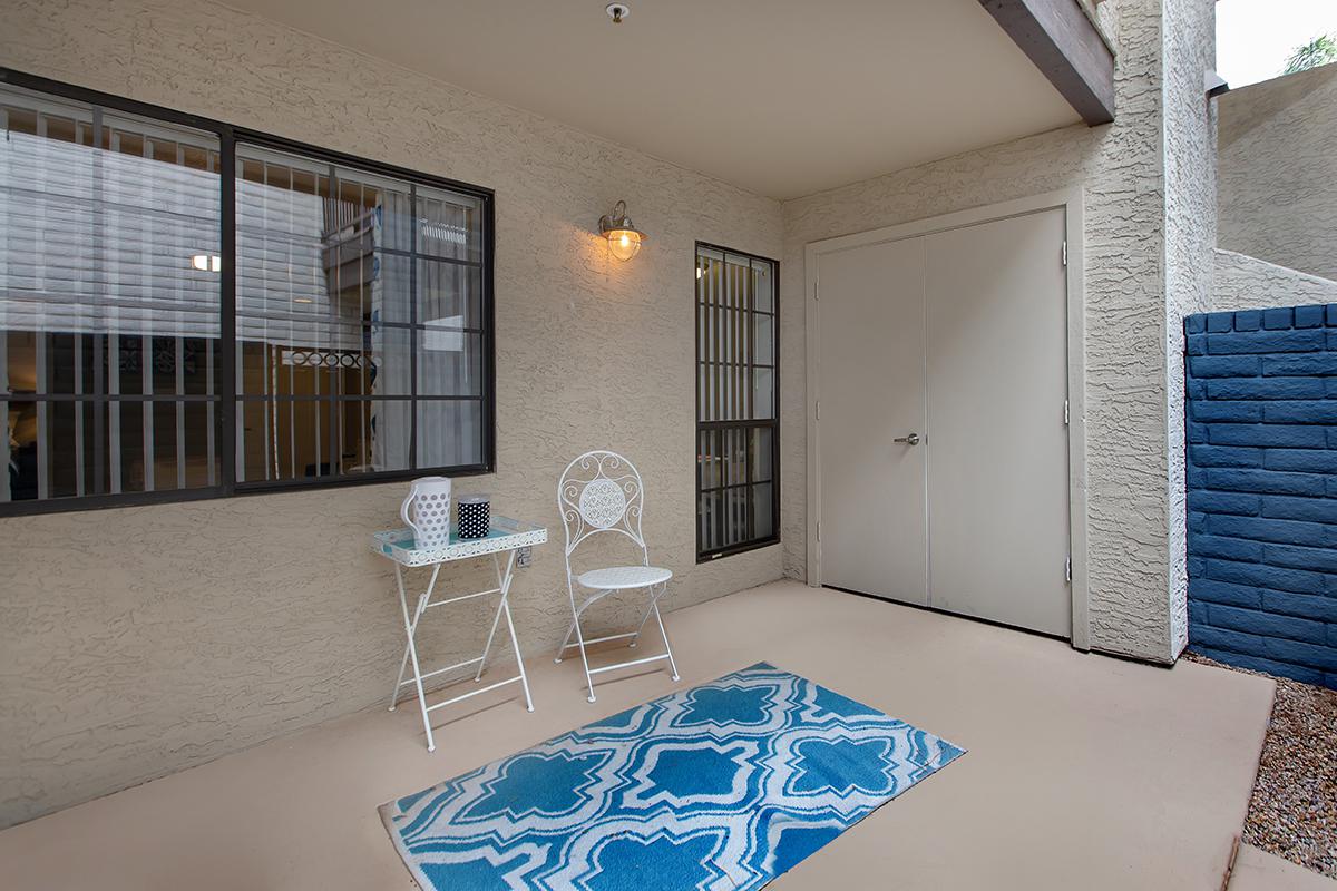 A small patio area with a light blue patterned rug on the ground. There is a white folding chair and a small table, next to a door leading inside. The walls are painted a neutral color, and there is a window with grid patterns nearby, adding to the cozy appearance of the space.