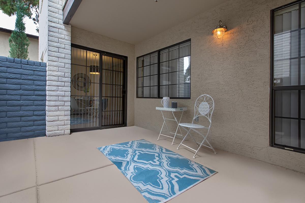 A small outdoor patio area featuring a light-colored floor, two white metal chairs, a small table, and a decorative blue patterned rug. There are two large windows with black frames, and a wall with a light fixture. A part of a garden wall can be seen in the background, adding greenery to the scene.