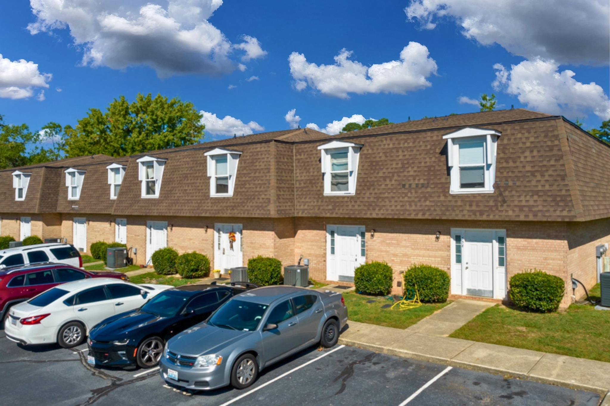 A row of suburban townhouses with sloped roofs and multiple windows, surrounded by well-maintained lawns. Several parked cars are visible in the foreground, and a clear blue sky with a few clouds is in the background.