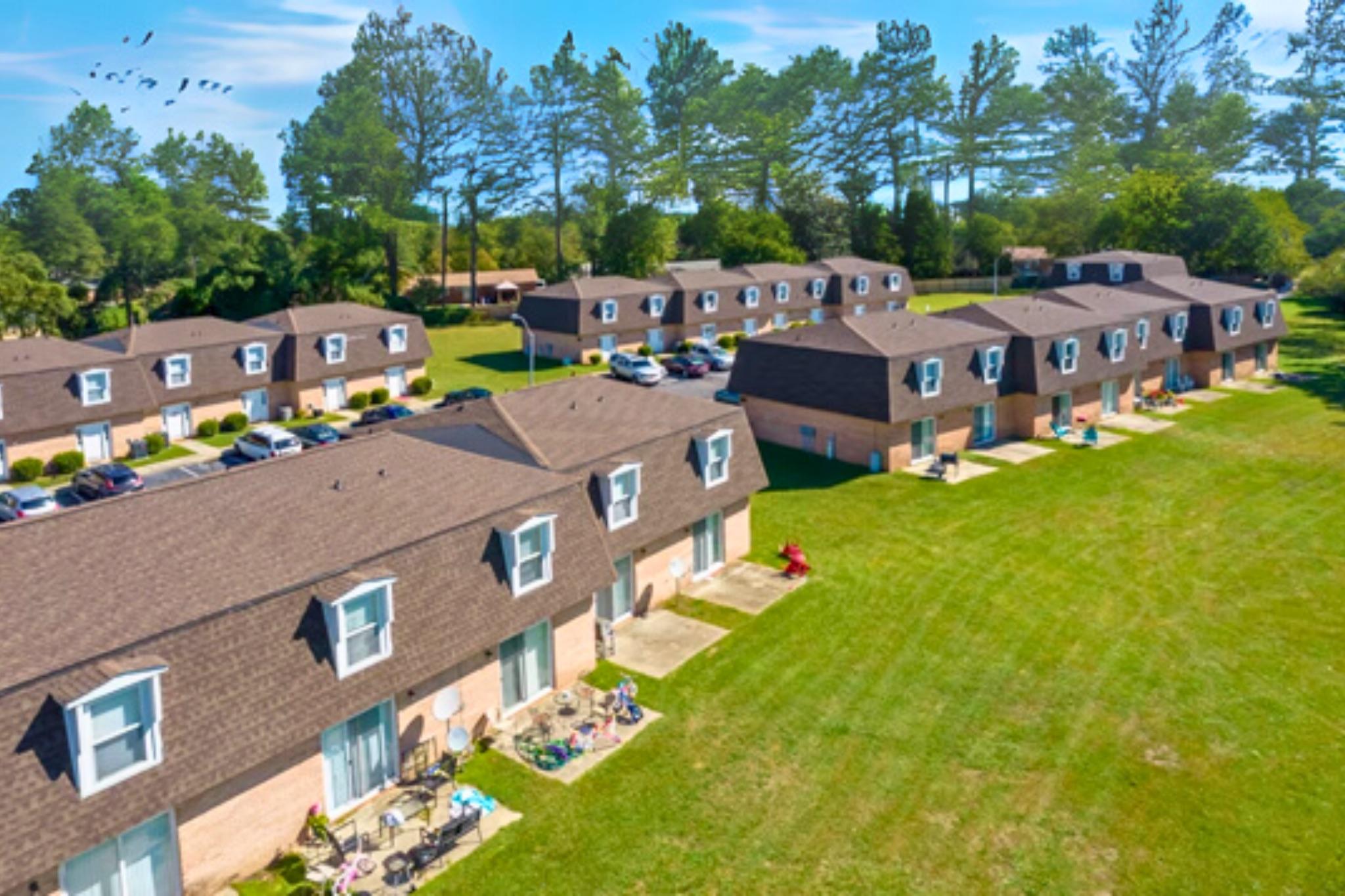 Aerial view of a residential complex featuring several small buildings with multiple units. Well-maintained lawns surround the structures, and parked cars are visible along the driveways. Tall trees line the background under a clear blue sky with a few birds flying in the distance.