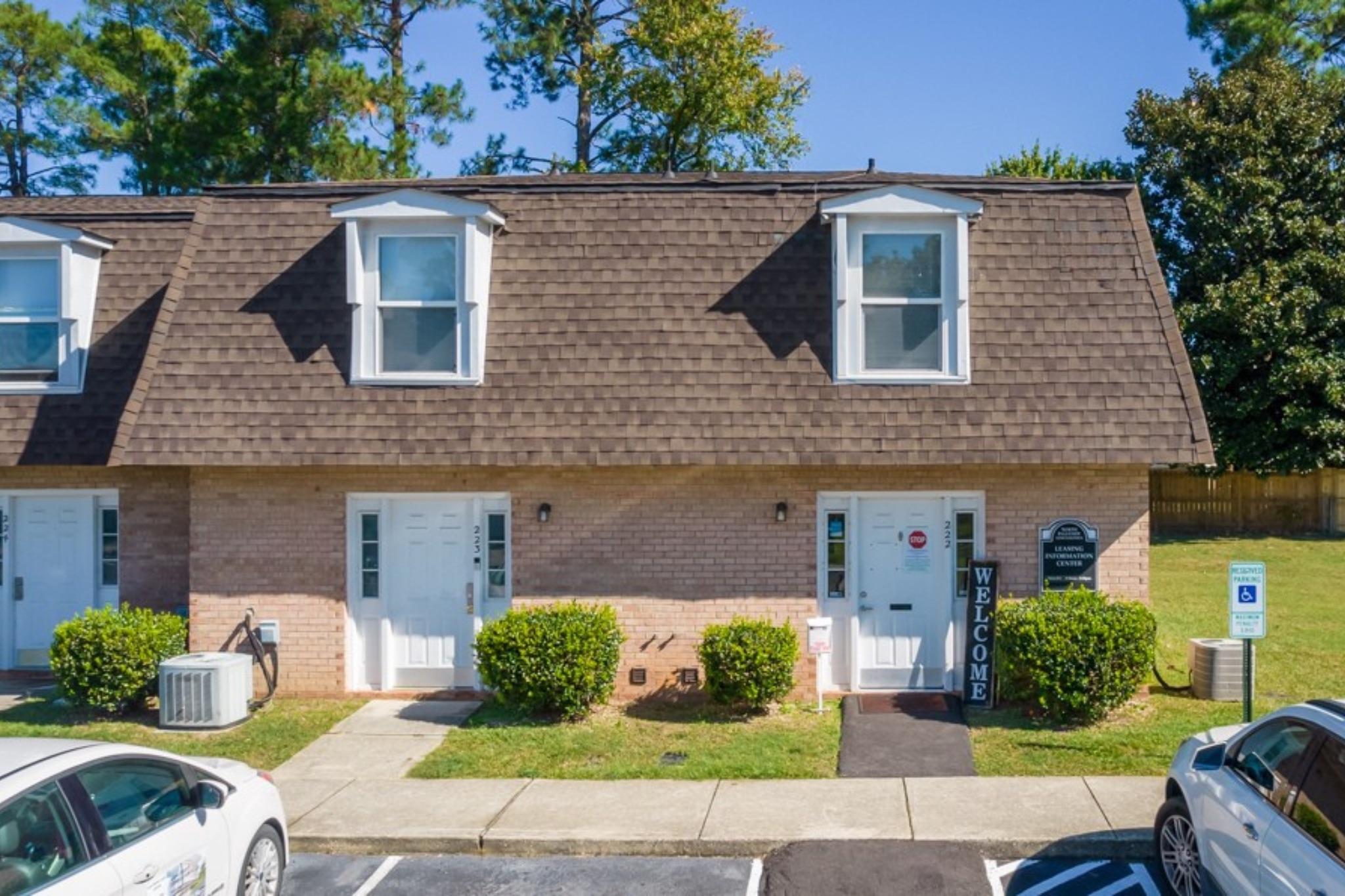 A two-story brick building with a brown shingled roof. The front has two doors, each with a white porch and small bushes on either side. A 'WELCOME' sign is displayed near the entrance. There are two parked cars in front of the building and a green grassy area. Trees are visible in the background under a clear blue sky.