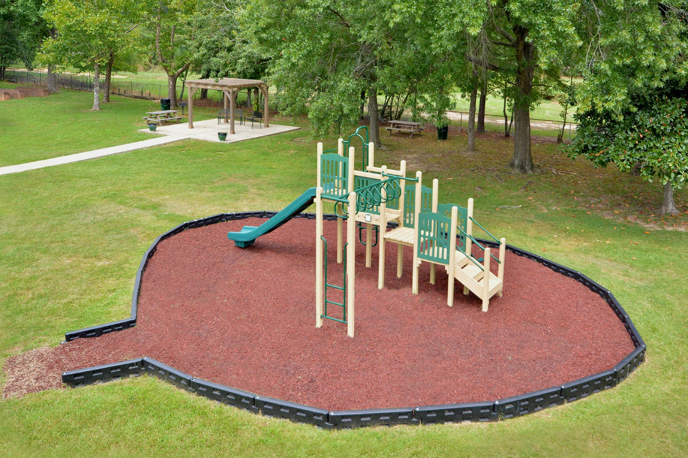A playground featuring a green slide, ladders, and climbing structures, situated on a bed of mulch. The playground is surrounded by grass, with trees in the background and a picnic area visible nearby.