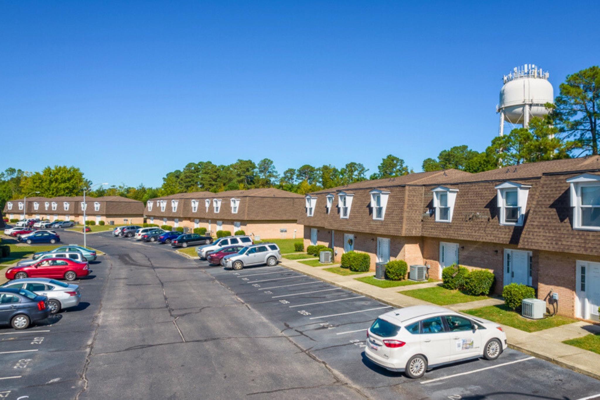 A residential area featuring multiple two-story apartment buildings with brown roofs. There are several parked cars along a paved lot, and grassy areas with shrubs. In the background, a water tower is visible against a clear blue sky. The setting appears well-maintained and suburban.