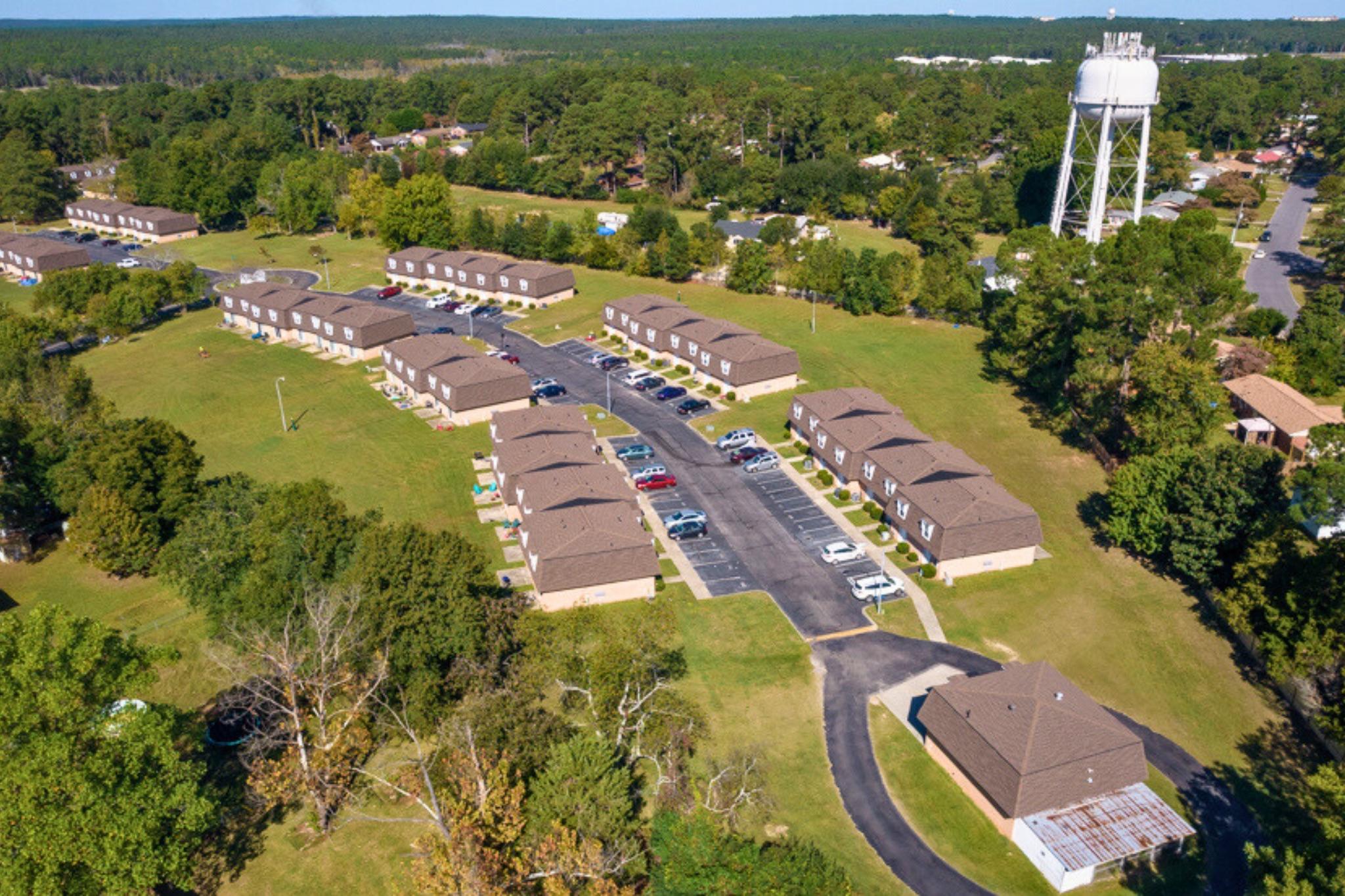 Aerial view of a residential area featuring several single-story apartment buildings arranged in rows. The scene includes well-maintained lawns, parked cars on a paved road, and a water tower in the background, surrounded by trees and greenery.