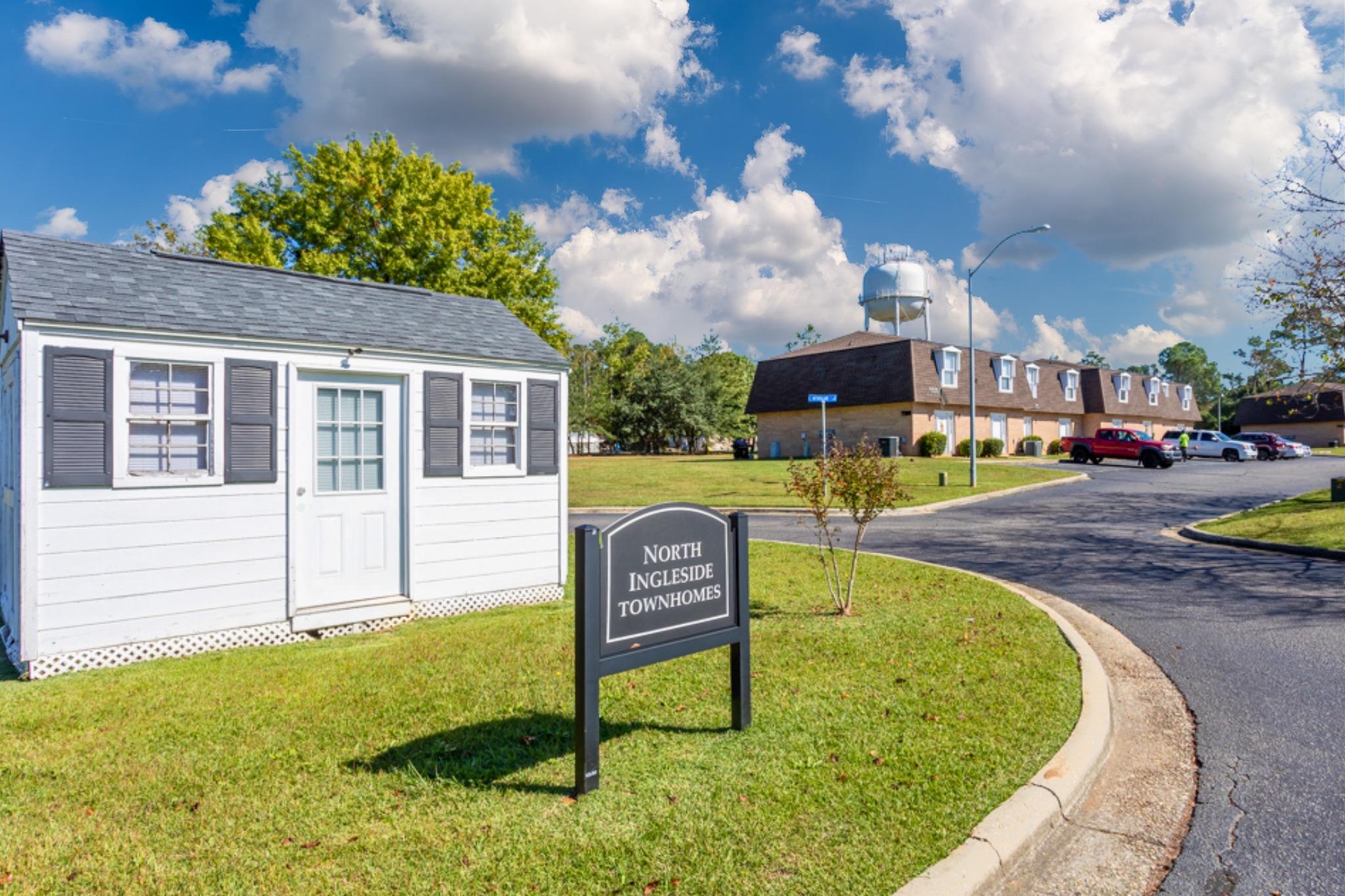 A view of North Ingleside Townhomes, featuring a white shed with a door and windows beside a sign displaying the name. In the background, there are two-story buildings and a water tower under a partly cloudy blue sky. The scene is well-maintained with green trees and landscaping.