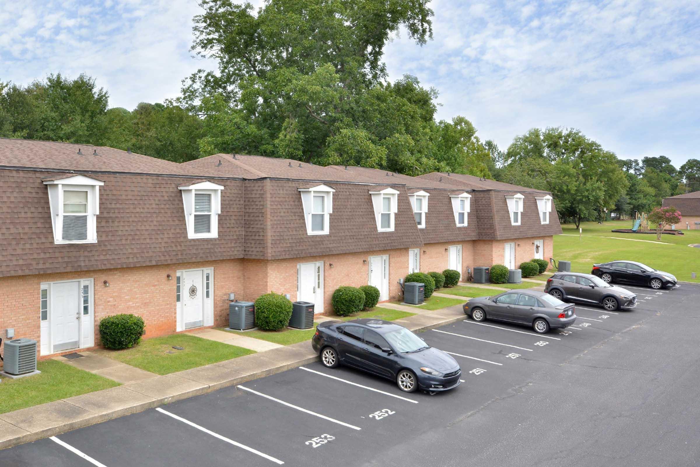 A row of brick apartment buildings with brown shingle roofs, featuring several parked cars in a well-maintained parking lot. The surrounding area includes green lawns and trees, under a partly cloudy sky.