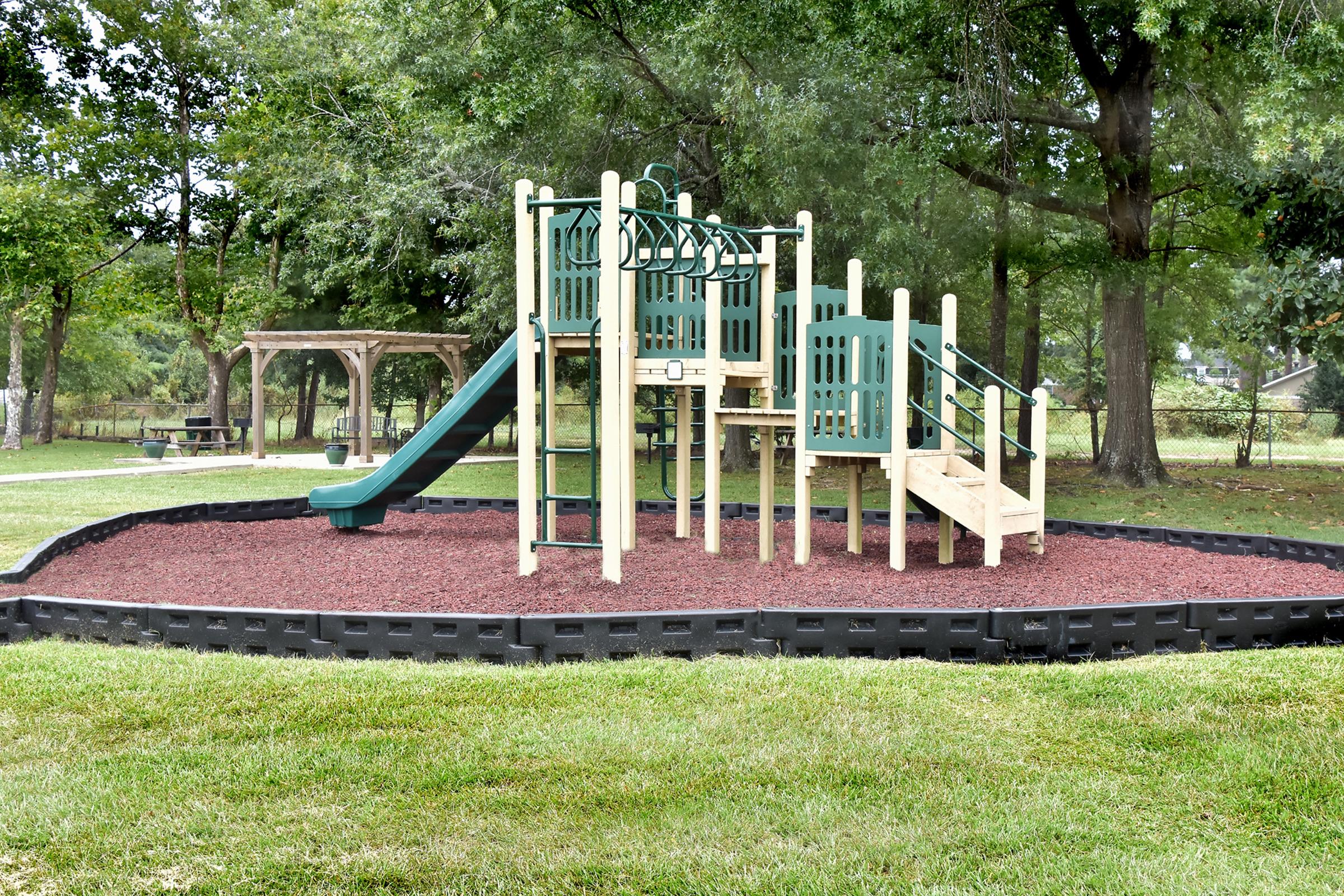 A playground structure featuring a slide, climbing features, and a ladder, surrounded by a rubber mulch safety surface. The area is bordered by a black plastic edging and set within a green space with trees in the background. Picnic tables are visible in the distance.