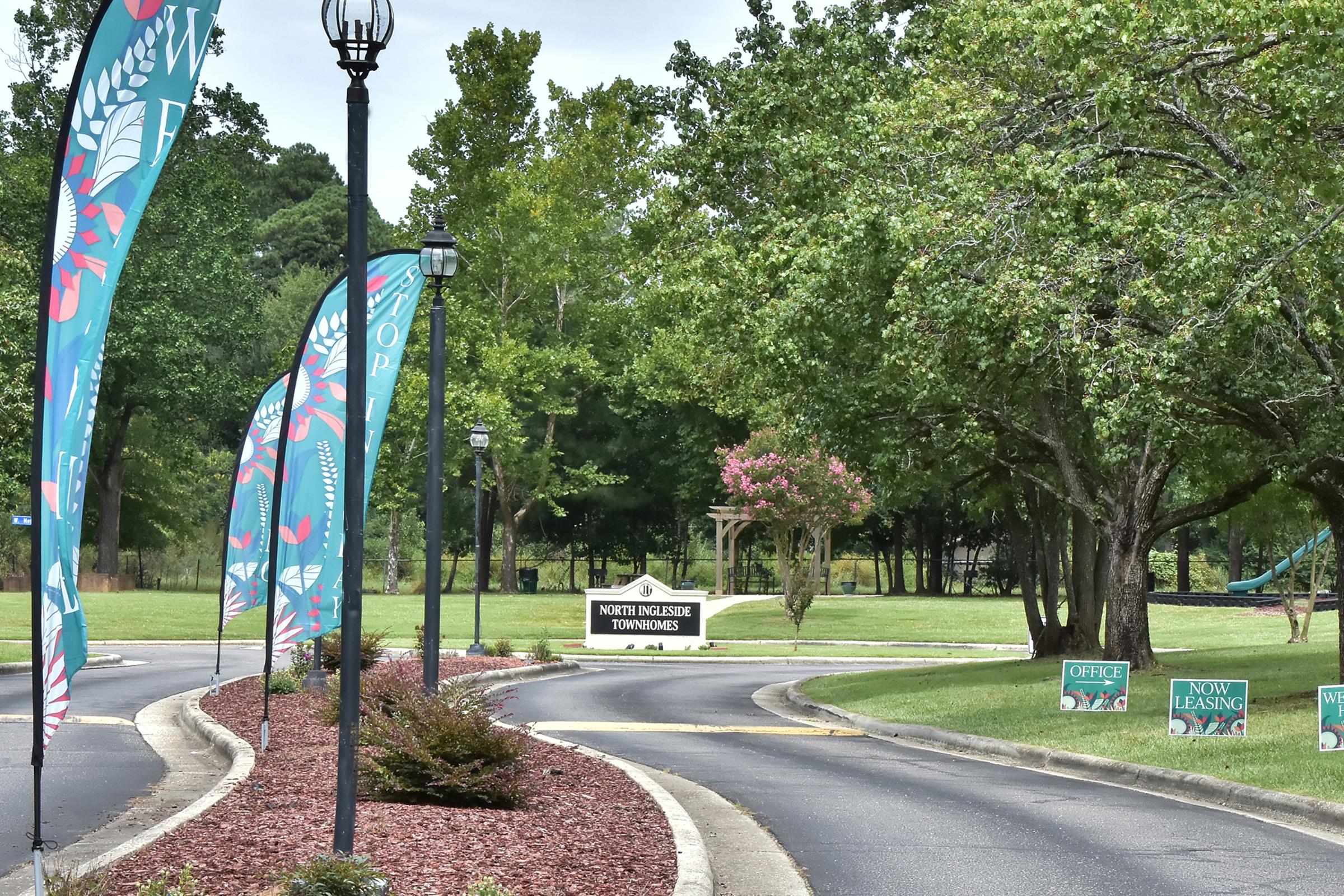 A winding path lined with colorful flags leads to a sign for "North Hillside Townhomes." Lush greenery and trees surround the area, with decorative lighting and leasing signs visible along the route. The scene conveys a welcoming atmosphere for potential residents.