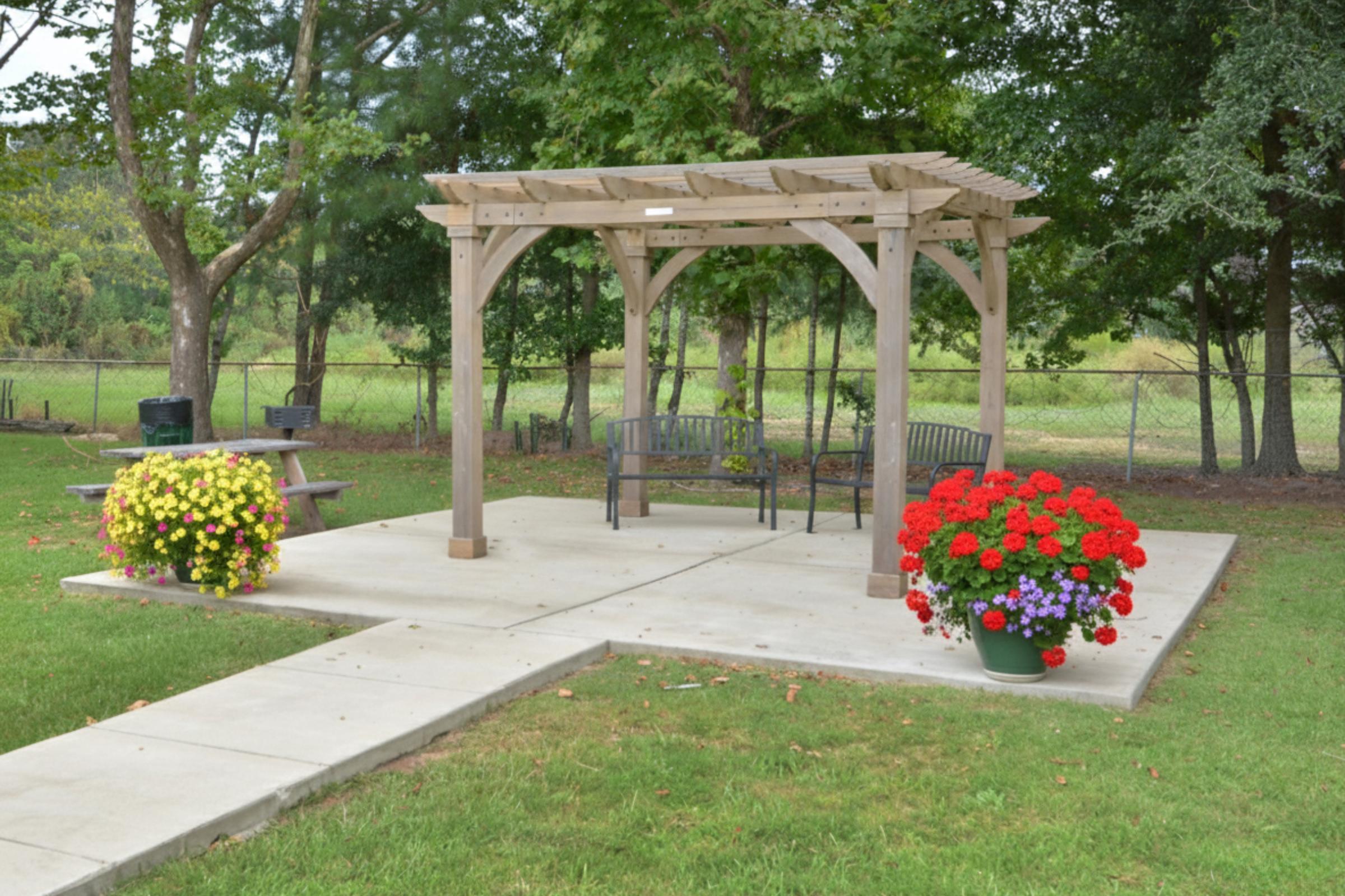 A wooden pergola with two benches is located on a concrete patio surrounded by lush green grass. Colorful flower arrangements in planters, including red and purple blooms, add vibrancy to the outdoor space, while trees and a fence form the backdrop.
