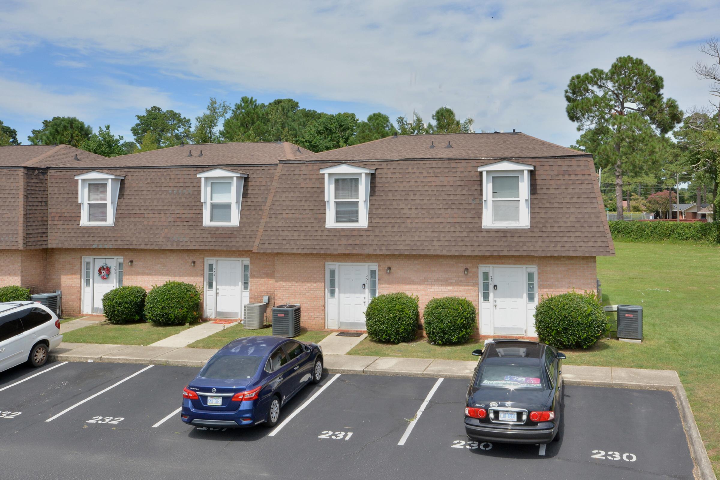 Two-story brick apartment building with a sloped roof and multiple units. Each unit has a white door and window. In the foreground, there are parked cars: a blue sedan and a darker-colored car. The surrounding area features green grass and trees, providing a landscaped appearance.