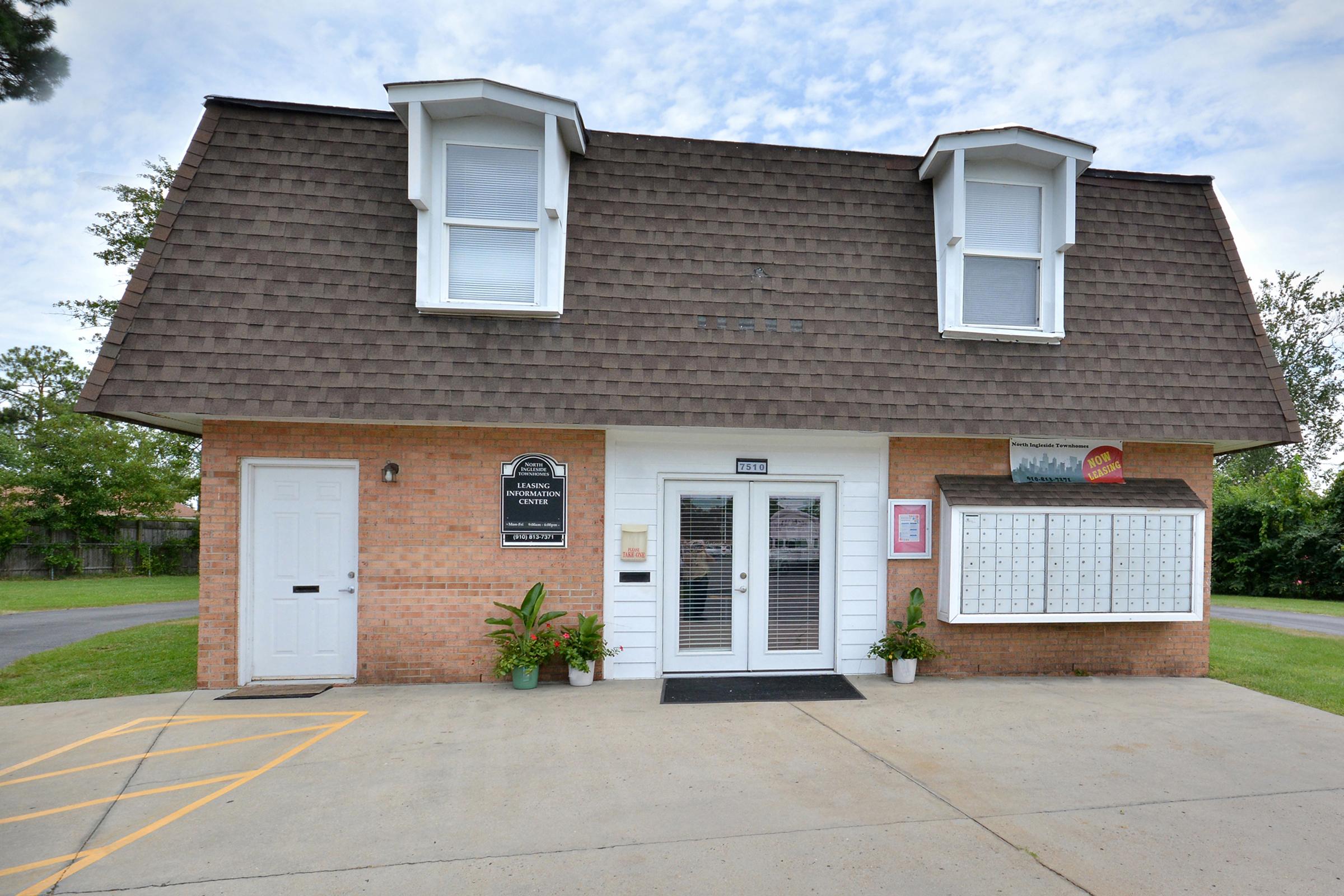 Two-story building with a brown shingle roof and white windows. The exterior is made of brick, featuring a sign for "Leasing Office." There are two doors, and a bulletin board next to a cluster of mailboxes. The area is surrounded by greenery and a clear blue sky.