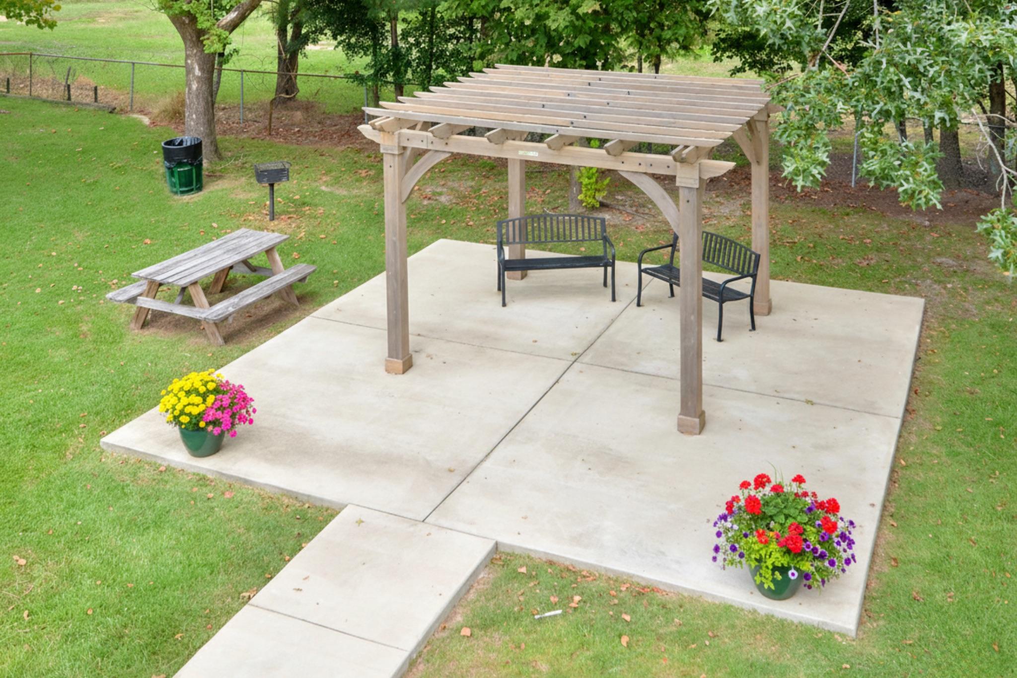 Aerial view of a landscaped outdoor area featuring a wooden pergola with two black benches, surrounded by vibrant flower pots. A picnic table is situated on a grassy section, with a concrete patio beneath the pergola. Lush trees border the scene, creating a serene and inviting atmosphere.
