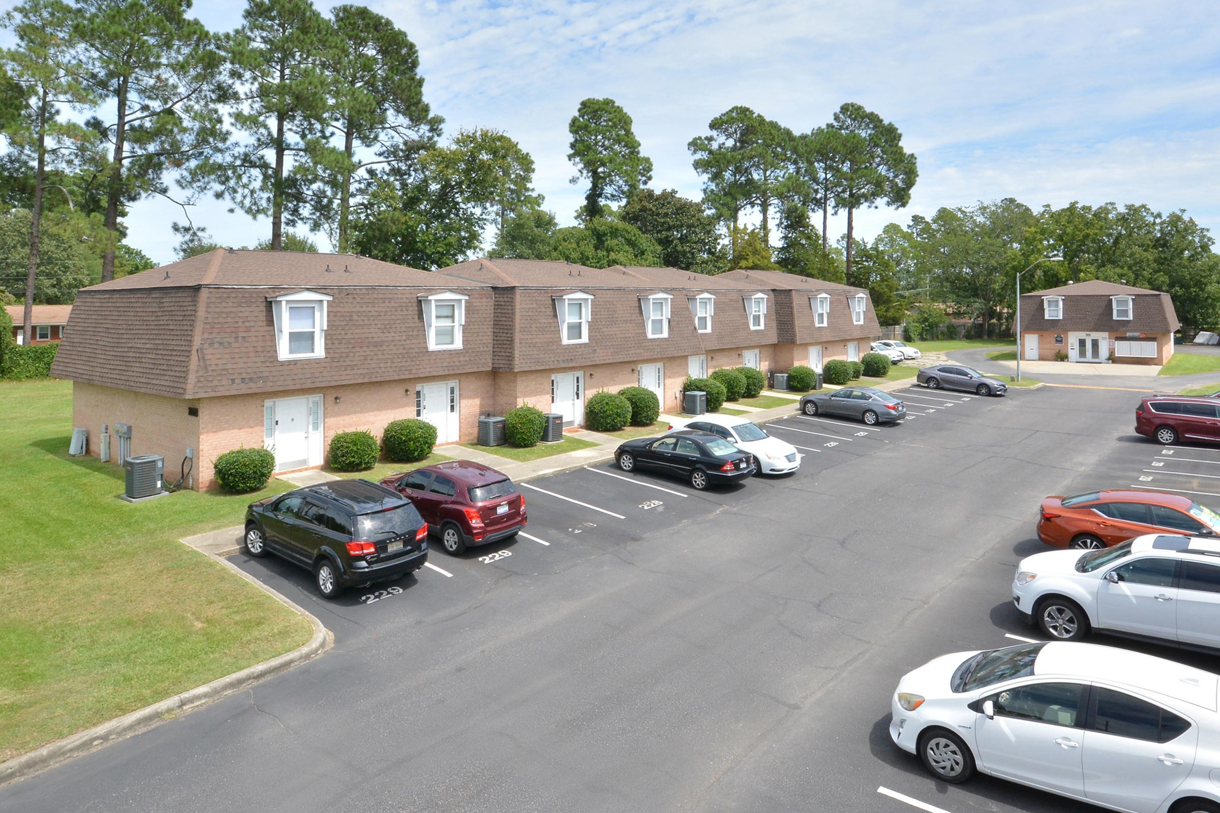 A view of a residential area featuring two-story brick townhouses with brown shingle roofs, surrounded by a well-maintained lawn. Several parked cars are visible in the foreground, and tall trees line the background under a partly cloudy sky.