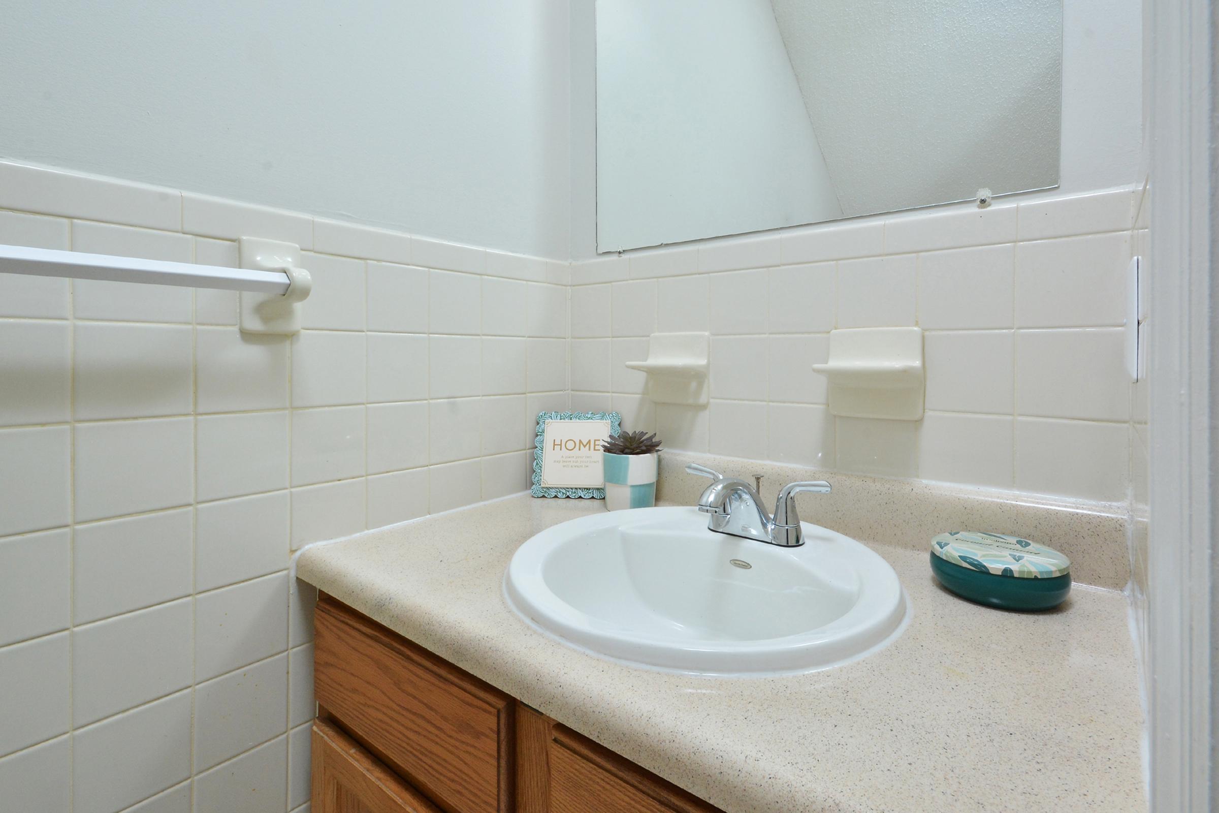 A clean bathroom sink area featuring a round sink, a faucet, and a countertop with wooden cabinetry. There are decorative items, including a small plant and a sign that says "Home." The walls are tiled, and a towel bar is visible. A mirror reflects the room's minimalist design.