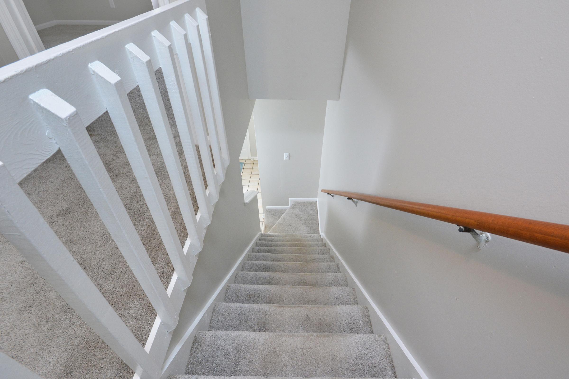 Staircase with beige carpet leading downwards, accompanied by a white railing on one side. The walls are painted light gray, and the bottom of the stairs opens to a room with tiled flooring. Soft natural light illuminates the space, enhancing the clean and modern aesthetic.