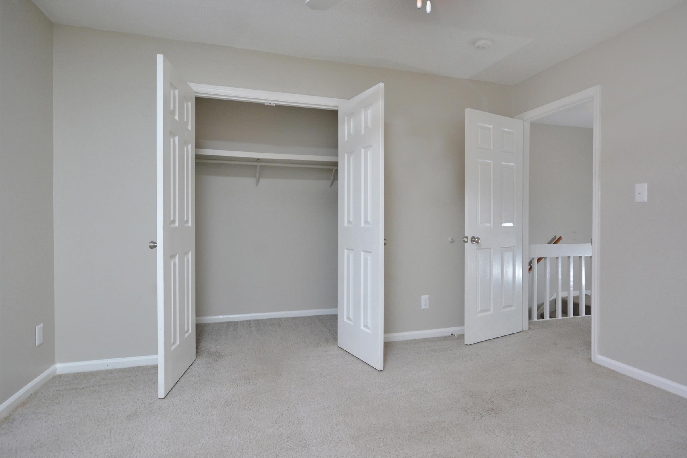 A clean, empty room with light gray walls and a light carpet. Two white doors are open, revealing a closet with no contents. A third door leads to a staircase. Natural light comes from a window, creating a bright and airy atmosphere. The room is spacious and uncluttered, suitable for various uses.