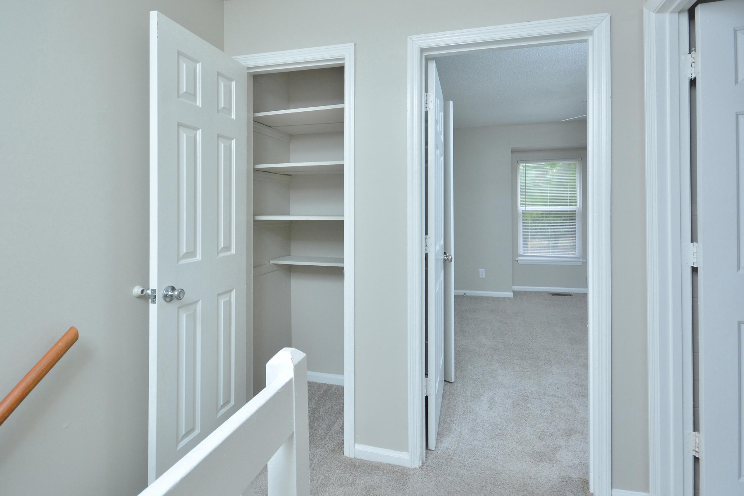 Interior view of a hallway featuring two doors open to separate rooms, with a built-in closet on the left. The walls are painted light gray, and the flooring is carpeted in a neutral color. Natural light enters through a window at the far end, creating a bright and spacious atmosphere.