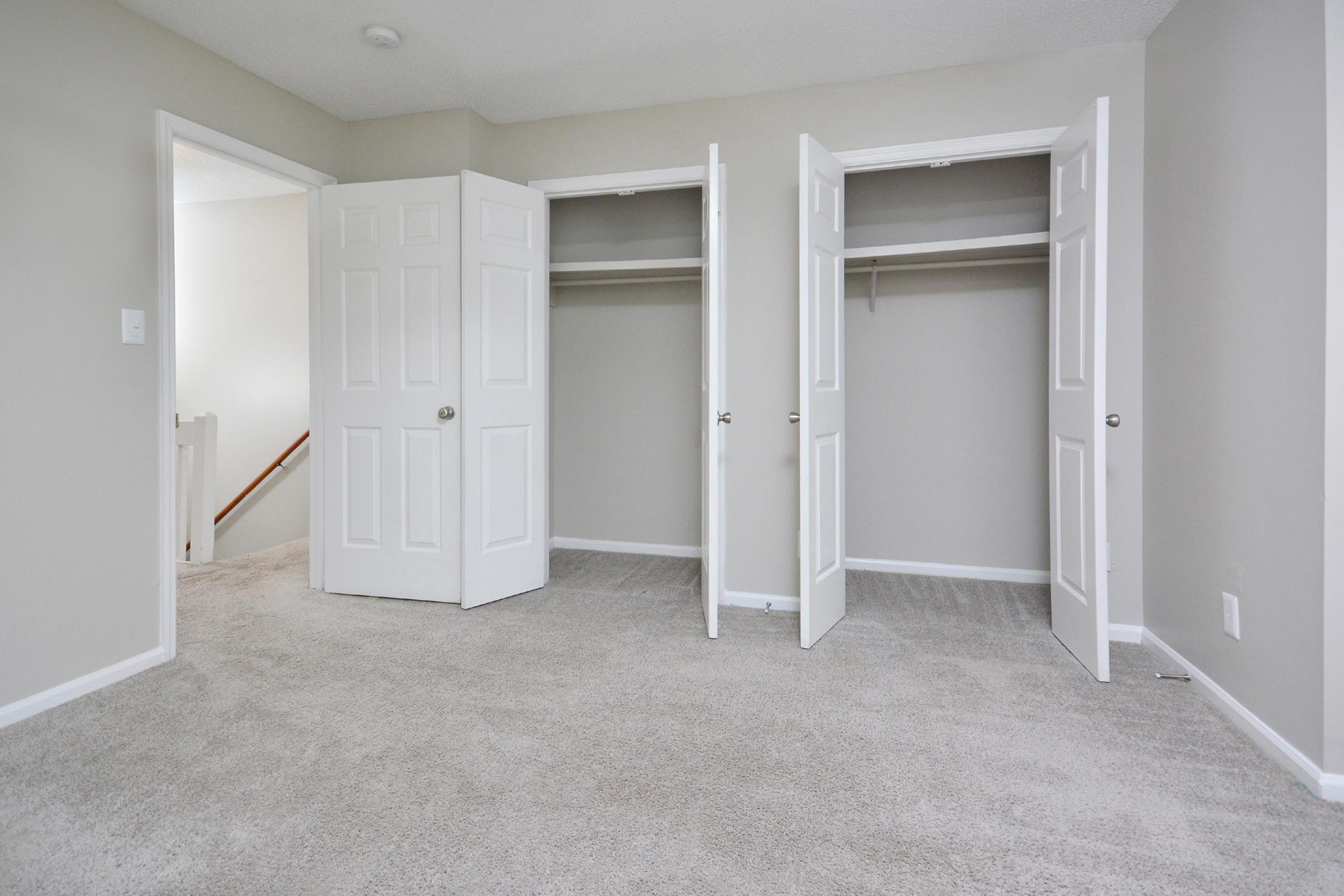 A spacious room featuring two open closets with white double doors. The walls are painted in a light color, and the floor is covered with beige carpet. In the background, a staircase is visible through an open door, suggesting access to another level. The overall atmosphere is bright and inviting.