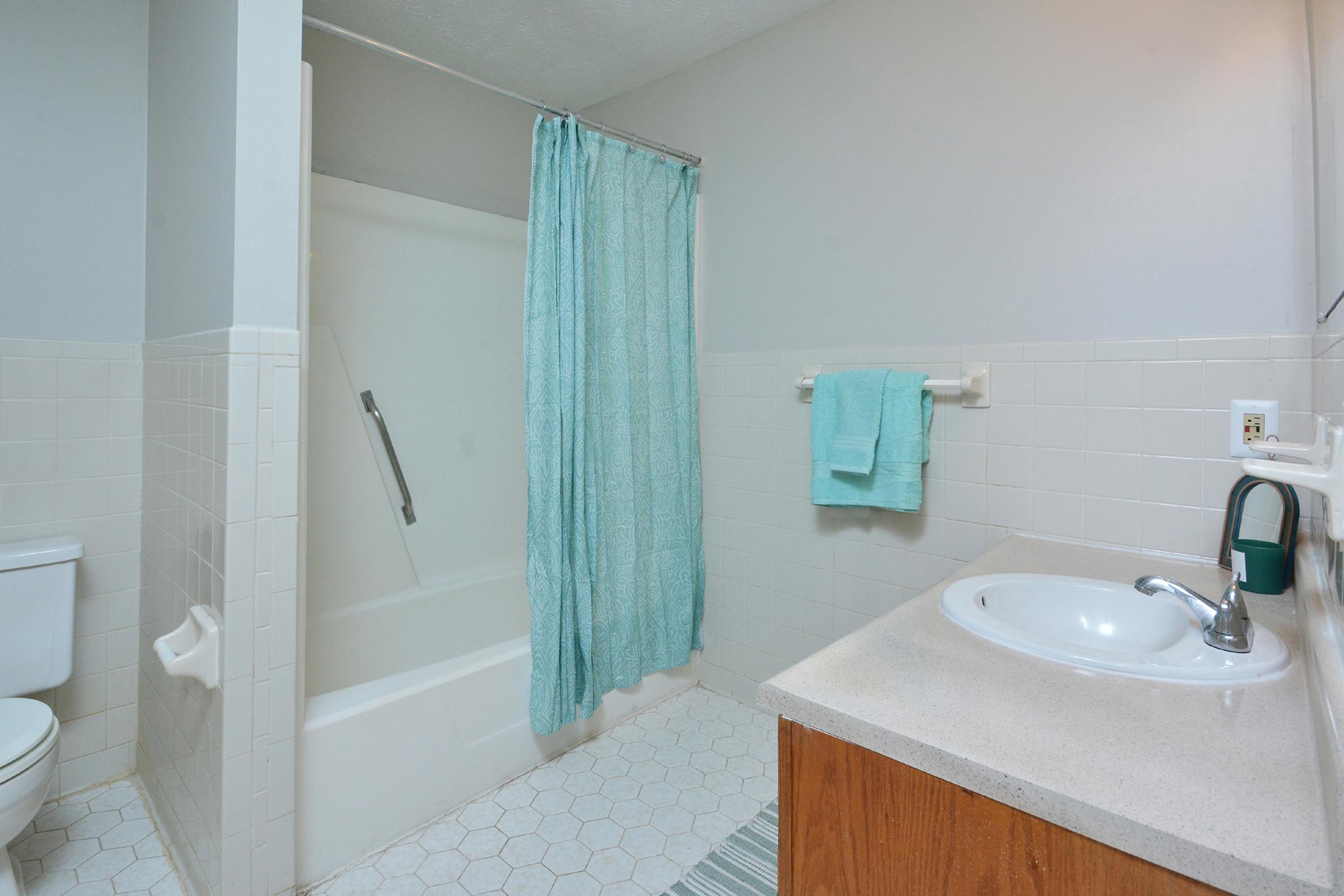 A clean, well-lit bathroom featuring a white bathtub with a light blue shower curtain, a white toilet, and a sink with a simple countertop. A light blue towel hangs on a rack and there is a small rug on the floor. The walls are painted in a soft, neutral color, creating a calming atmosphere.