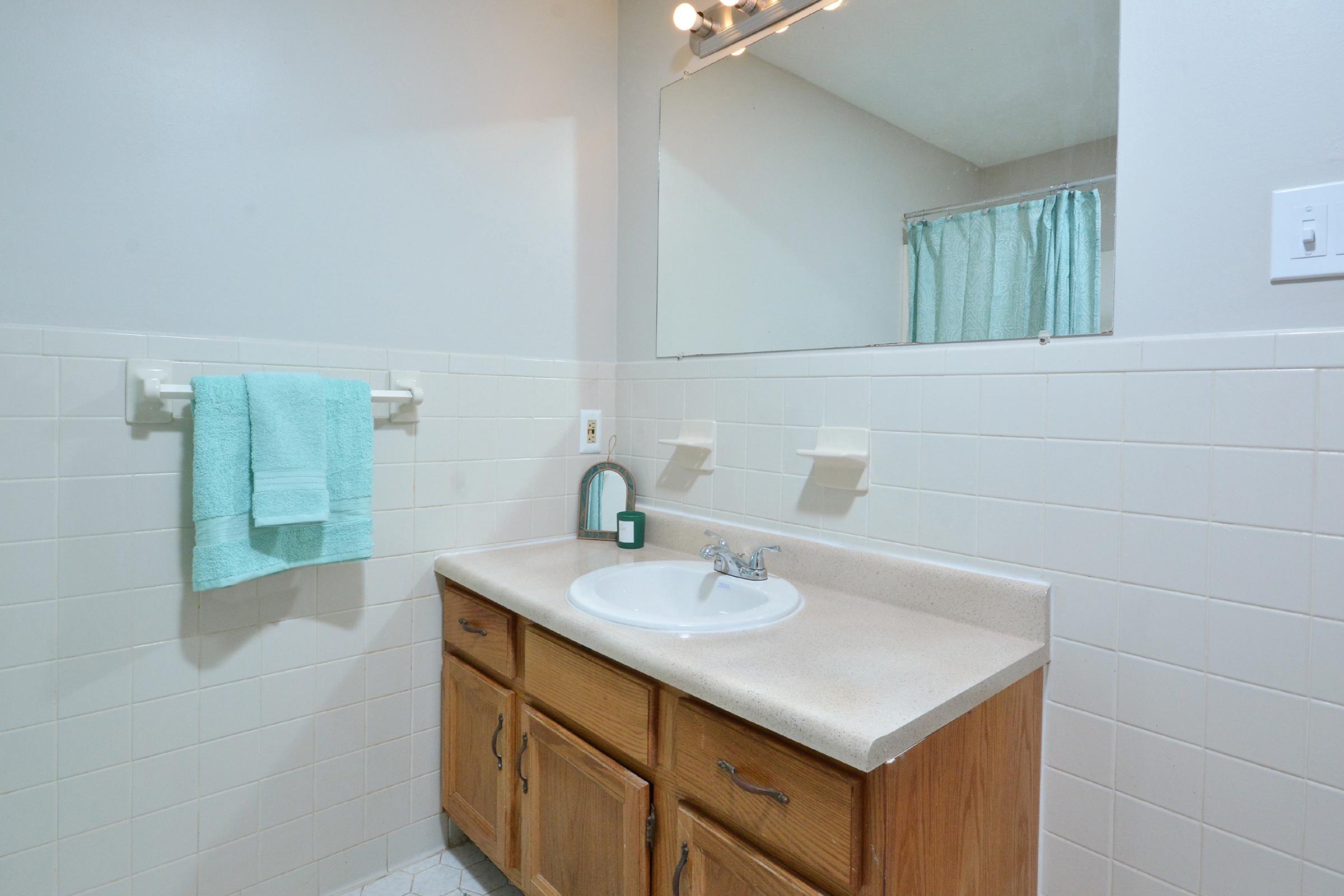 A clean bathroom featuring a light-colored vanity with a sink, a mirror above, and two towel holders. There are two aqua-colored towels hanging on the wall, and a light blue shower curtain is partially visible in the background. The walls are tiled in a light shade, creating a bright and airy atmosphere.