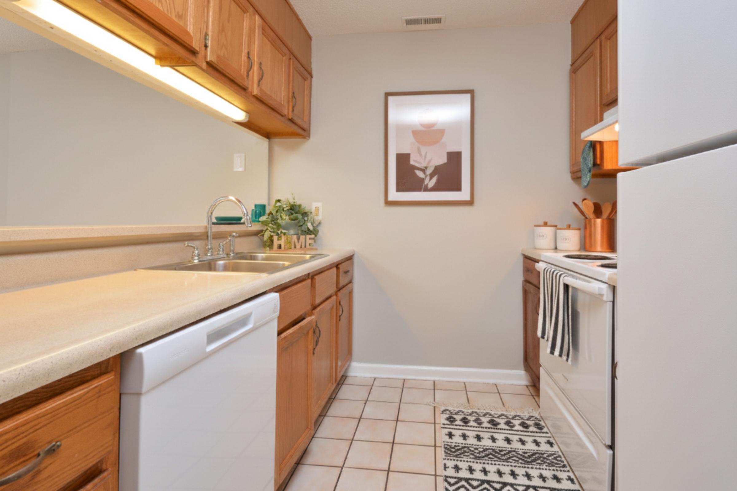 A well-organized kitchen featuring wooden cabinets, a double sink, and modern appliances. The space includes a refrigerator, stove, and dishwasher, with a decorative print on the wall. A stylish runner rug adds charm to the tiled floor, creating a warm and inviting atmosphere.