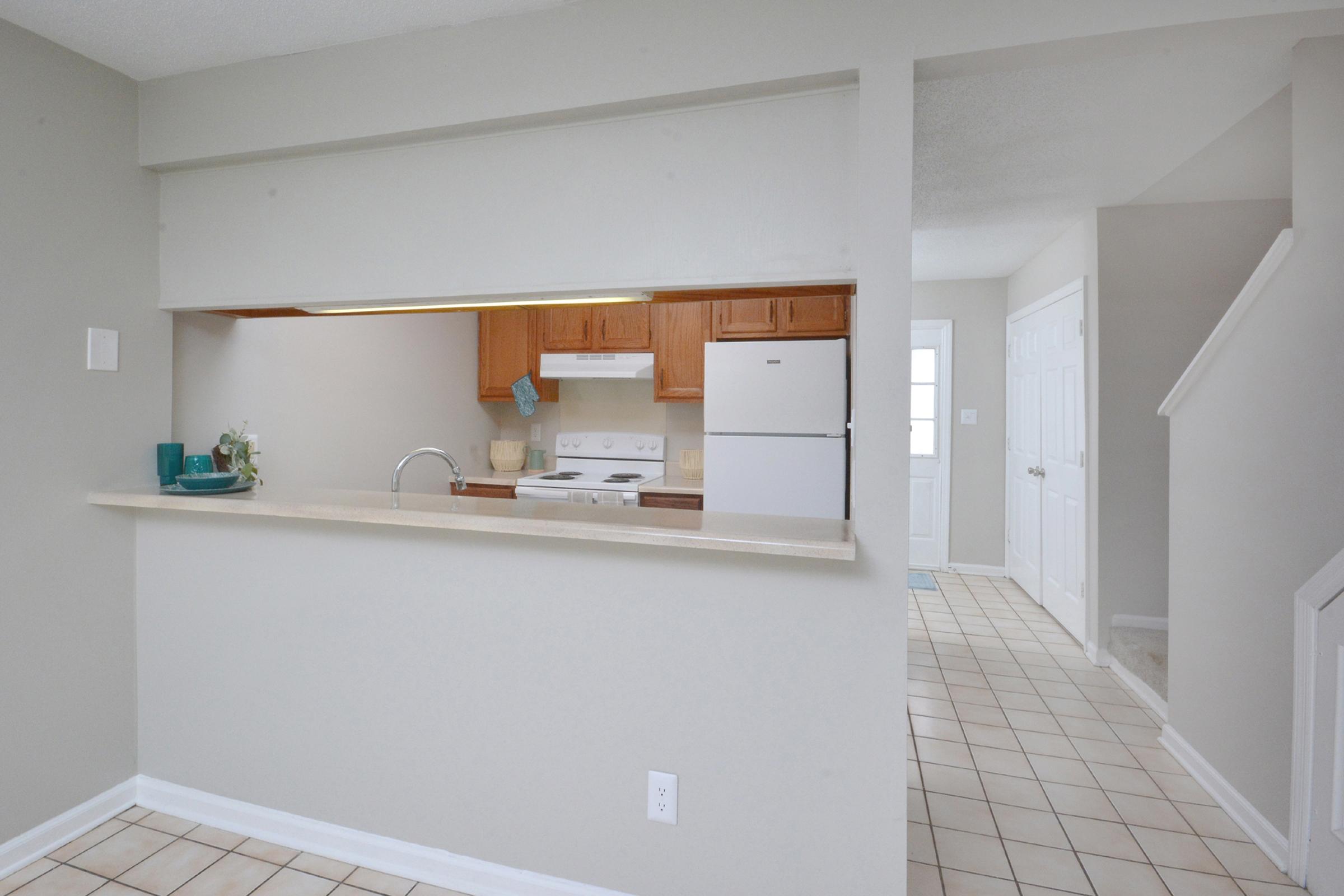 A view of a modern kitchen from an open-plan living area. The kitchen features wooden cabinets, a white stove, and refrigerator, with a countertop separating it from the living space. Natural light enters through a nearby door, illuminating the tiled floor and light gray walls.