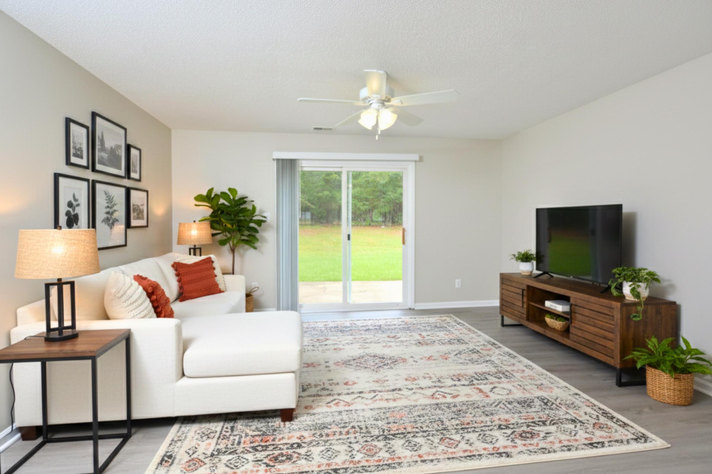 A bright, cozy living room featuring a white sofa with orange accents, a wooden TV stand, and a decorative rug. The space has a sliding glass door leading to a backyard view, with greenery outside. Two lamps and potted plants add warmth and style to the room's decor.