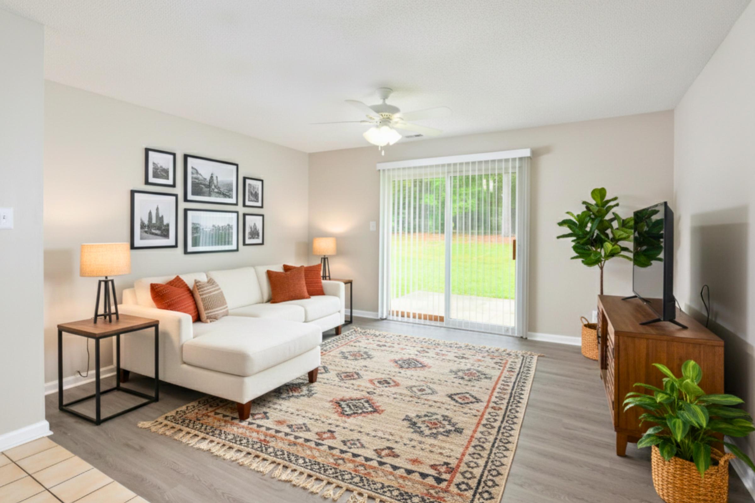 A cozy living room featuring a light-colored sectional sofa with orange and beige cushions, framed black-and-white photographs on the wall, a wooden TV stand with a television, and a potted plant. Sliding glass doors lead to a grassy outdoor area, with a decorative area rug on the floor.