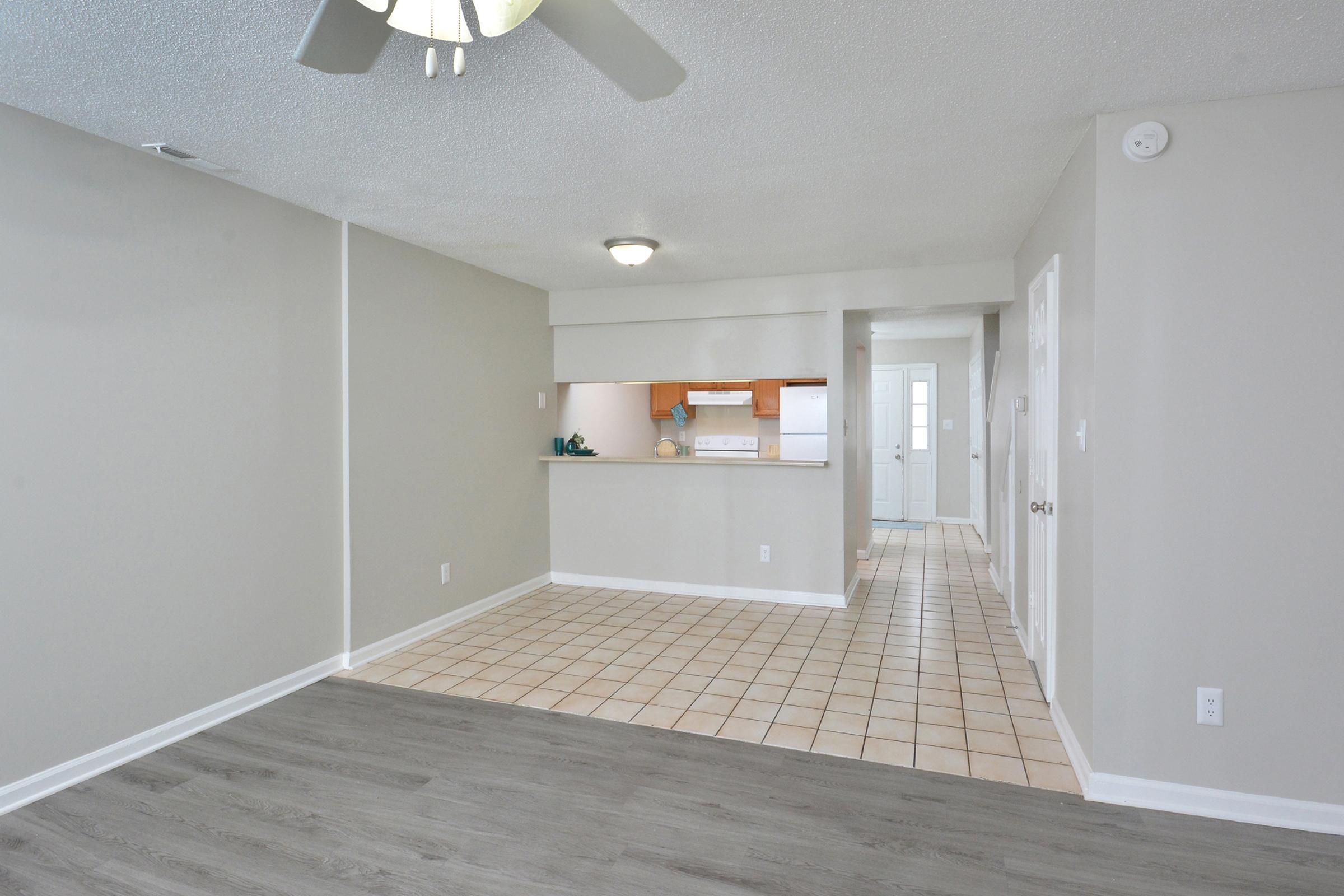Interior of an apartment featuring a living area with light gray walls and a wood-style floor. A kitchen with a tiled floor is visible in the background, showcasing an open layout. There is a ceiling fan in the living room area, and the space is well-lit with natural light coming from an entrance in the back.