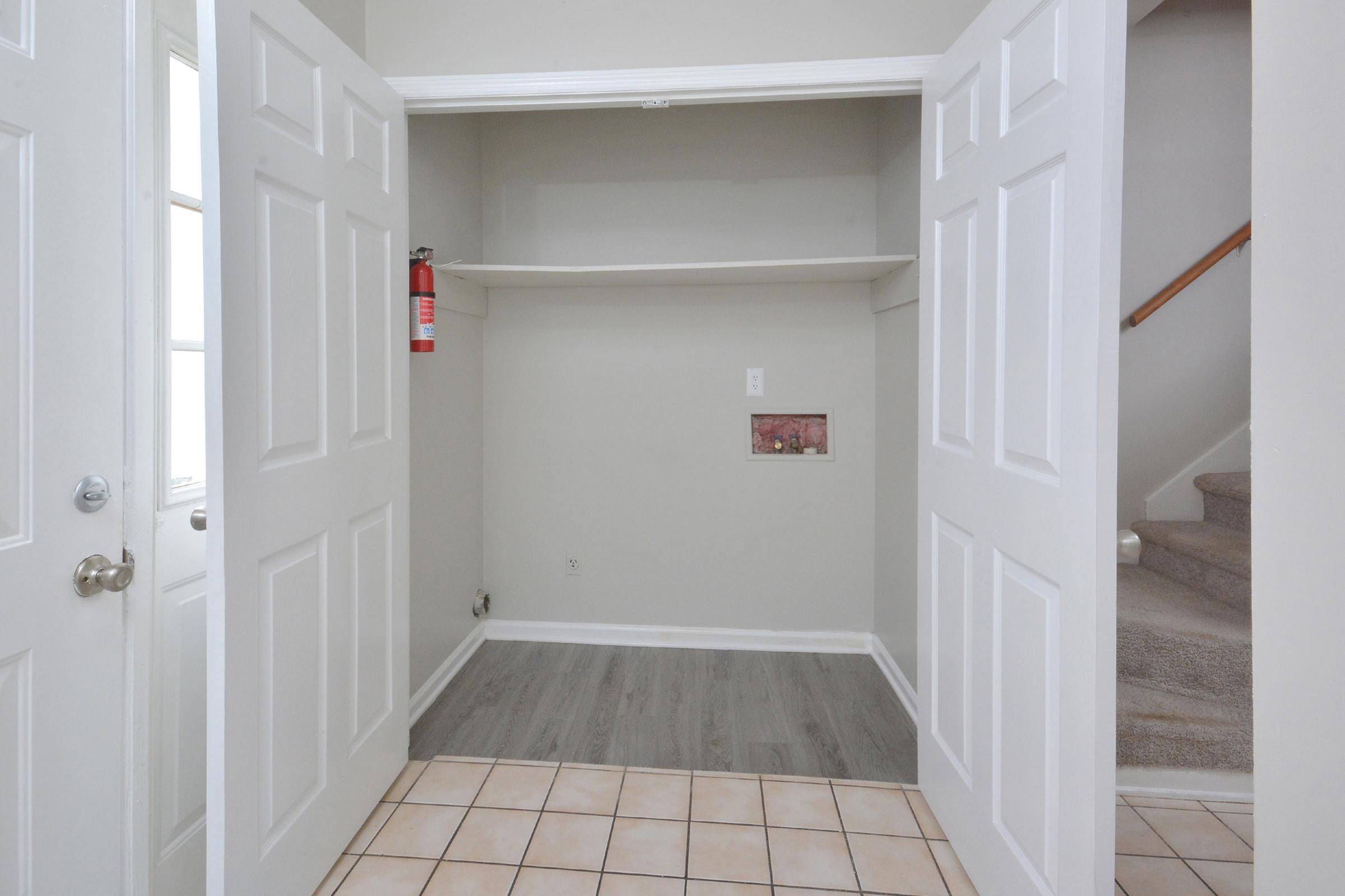 A view of an empty closet with two white doors open. The interior features a shelf, a small wall panel, and light-colored walls. The floor has tiling transitioning to a wood-like laminate. A staircase is visible in the background, leading to a second level. A fire extinguisher is mounted on the wall.