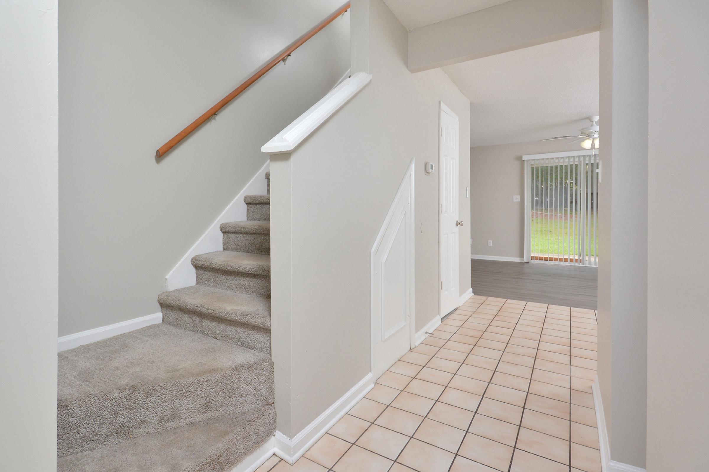 A view of an interior space showcasing a staircase with carpeted steps on the left, leading to a second level. The walls are light-colored, and the flooring is a mix of tile and carpet. To the right, there is a hallway leading to a bright, open area with sliding glass doors.