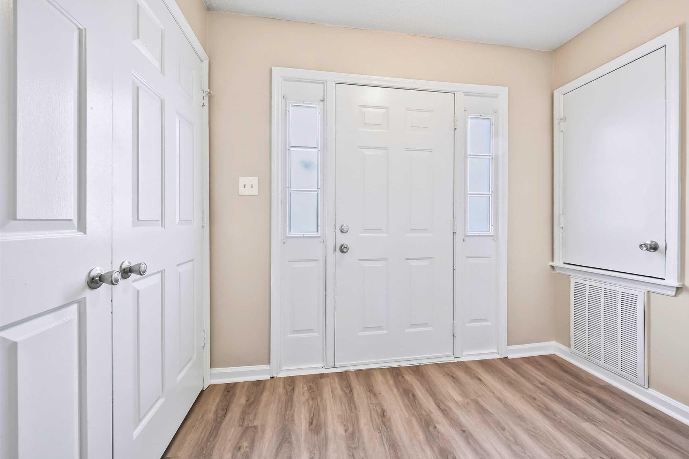 A clean and neutral entryway featuring a set of double white doors with glass panels. Light-colored walls complement the wooden floor. Two closet doors are visible on the left, and a small wall vent is located on the right. The space is well-lit and inviting.