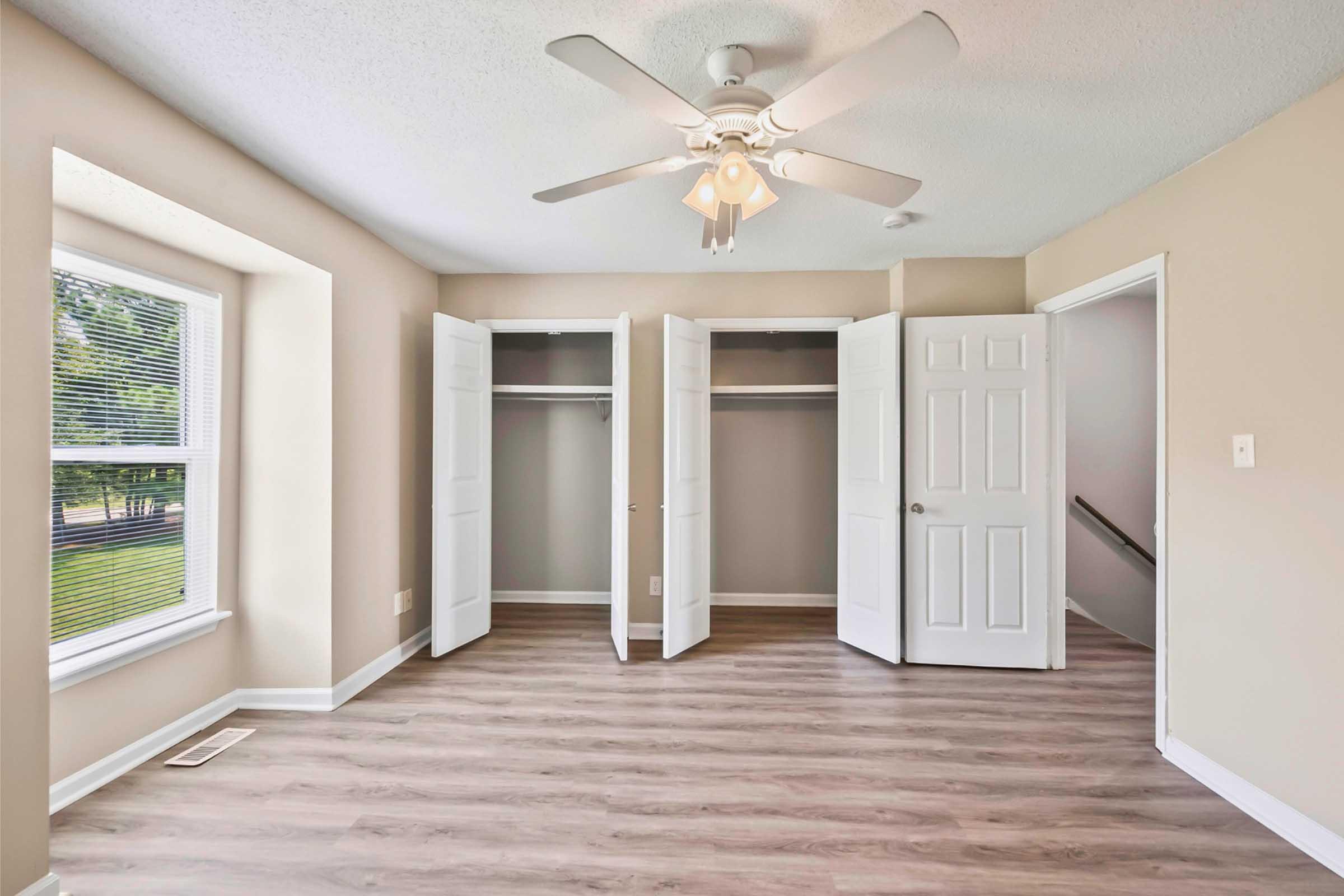 A well-lit bedroom featuring light-colored walls, a ceiling fan with light fixture, and a large window. There are two sets of double doors leading to closets, and the floor is covered with laminate wood flooring. The room is empty, providing a spacious and open feel.