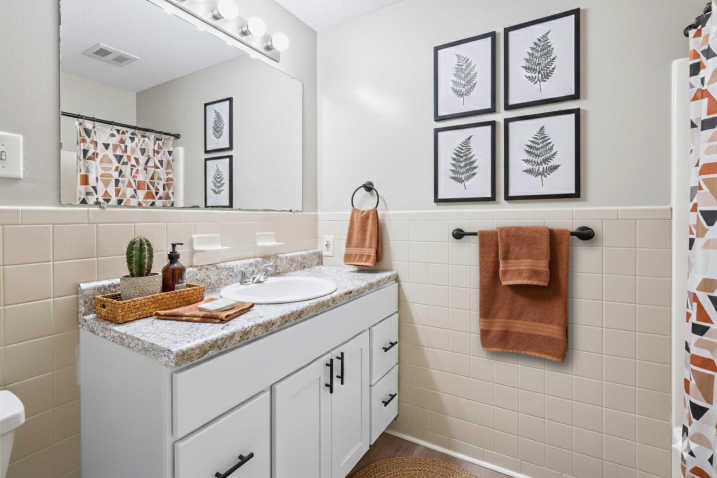 A modern bathroom featuring a white vanity with a sink, a large mirror with vanity lights, and decorative fern prints on the wall. There are two brown towels hanging, a small basket with toiletries, and a cactus plant on the countertop. The walls are painted light gray, and the shower curtain has a geometric pattern.