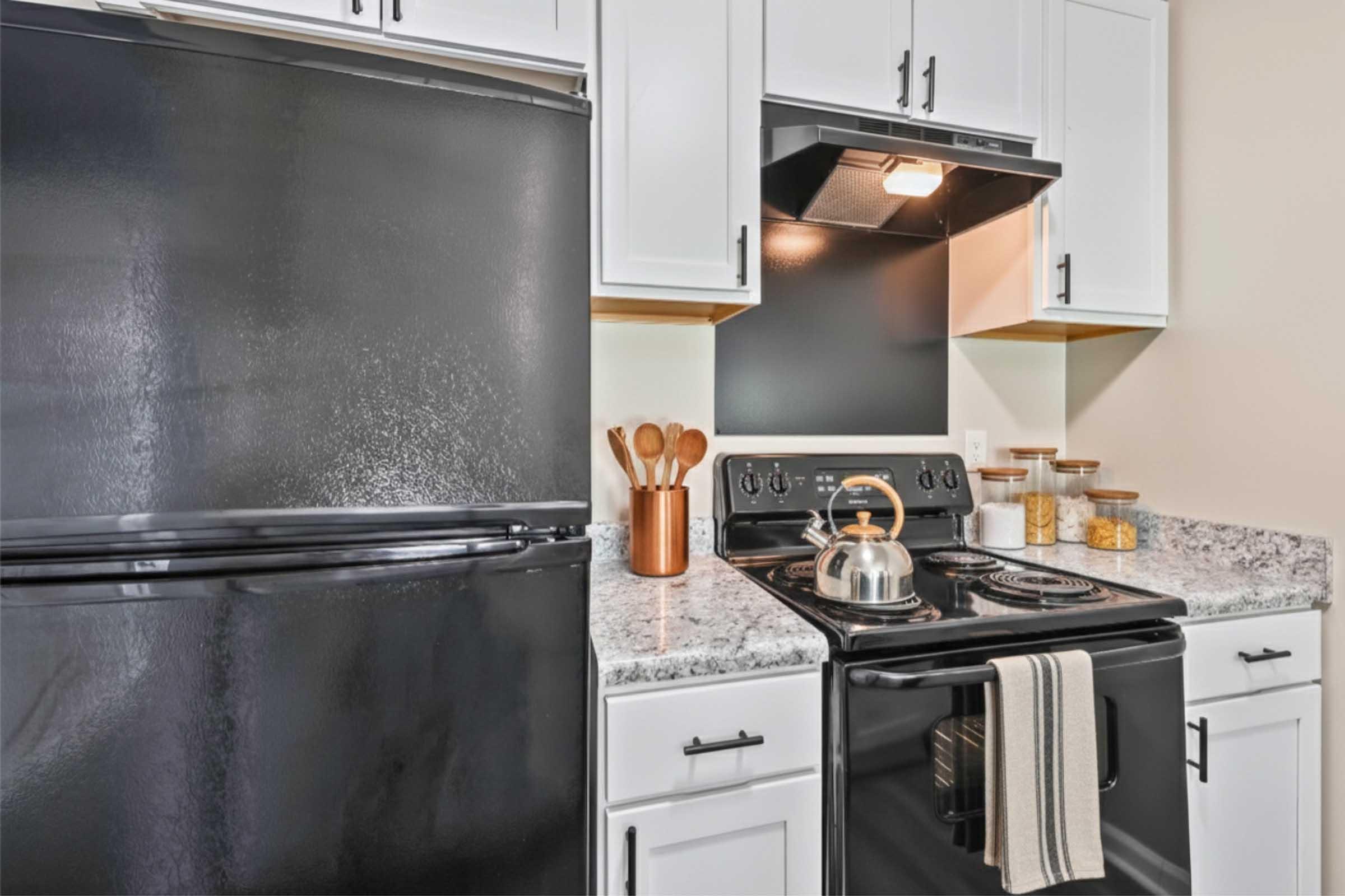 A modern kitchen featuring a black refrigerator, a black stove with an overhead range hood, and a countertop made of granite. Wooden utensils are displayed in a copper holder, complemented by jars of ingredients in the background. The cabinetry is white, adding brightness to the space.