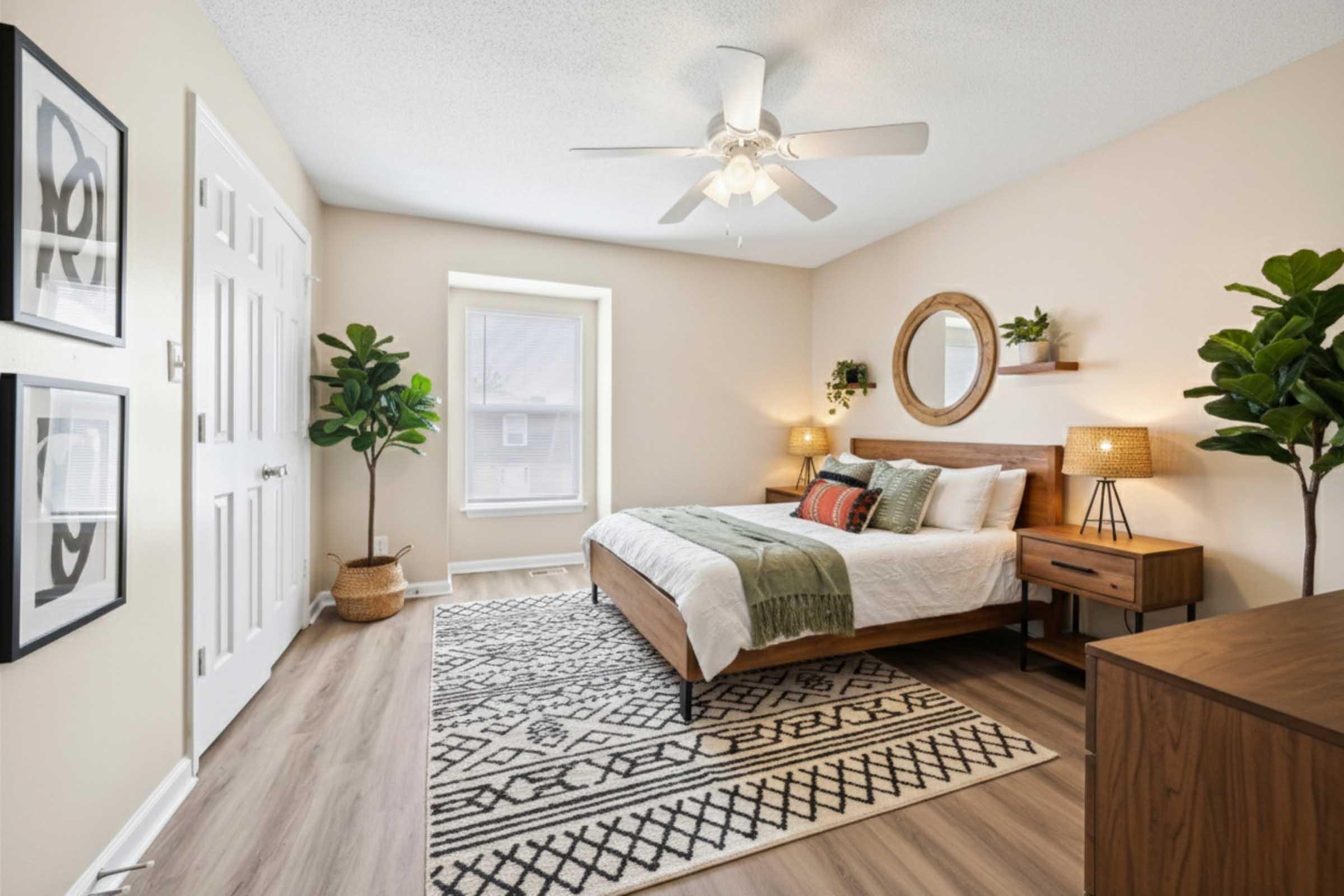 A cozy bedroom featuring a wooden bed with decorative pillows, two bedside lamps, and a round mirror above the bed. A large window allows natural light. The room includes a patterned area rug, a dresser, and potted plants, creating a welcoming and contemporary atmosphere.