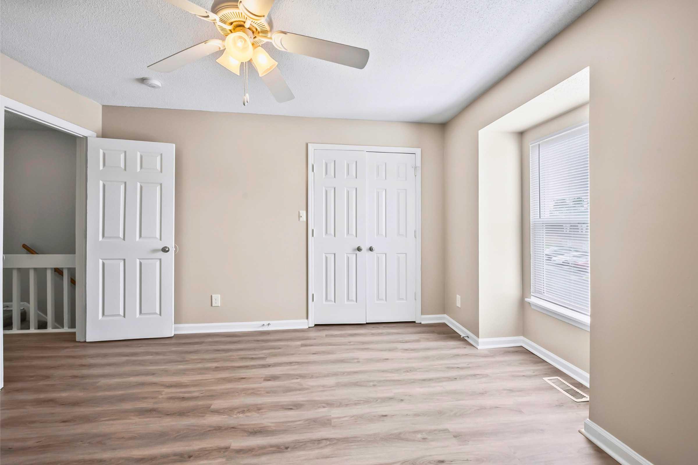 Empty room featuring light-colored walls, a ceiling fan, and laminate flooring. There are two closed double doors on the left, leading to a closet or storage area, and a window with blinds on the right allowing natural light. The space appears clean and ready for furnishing.