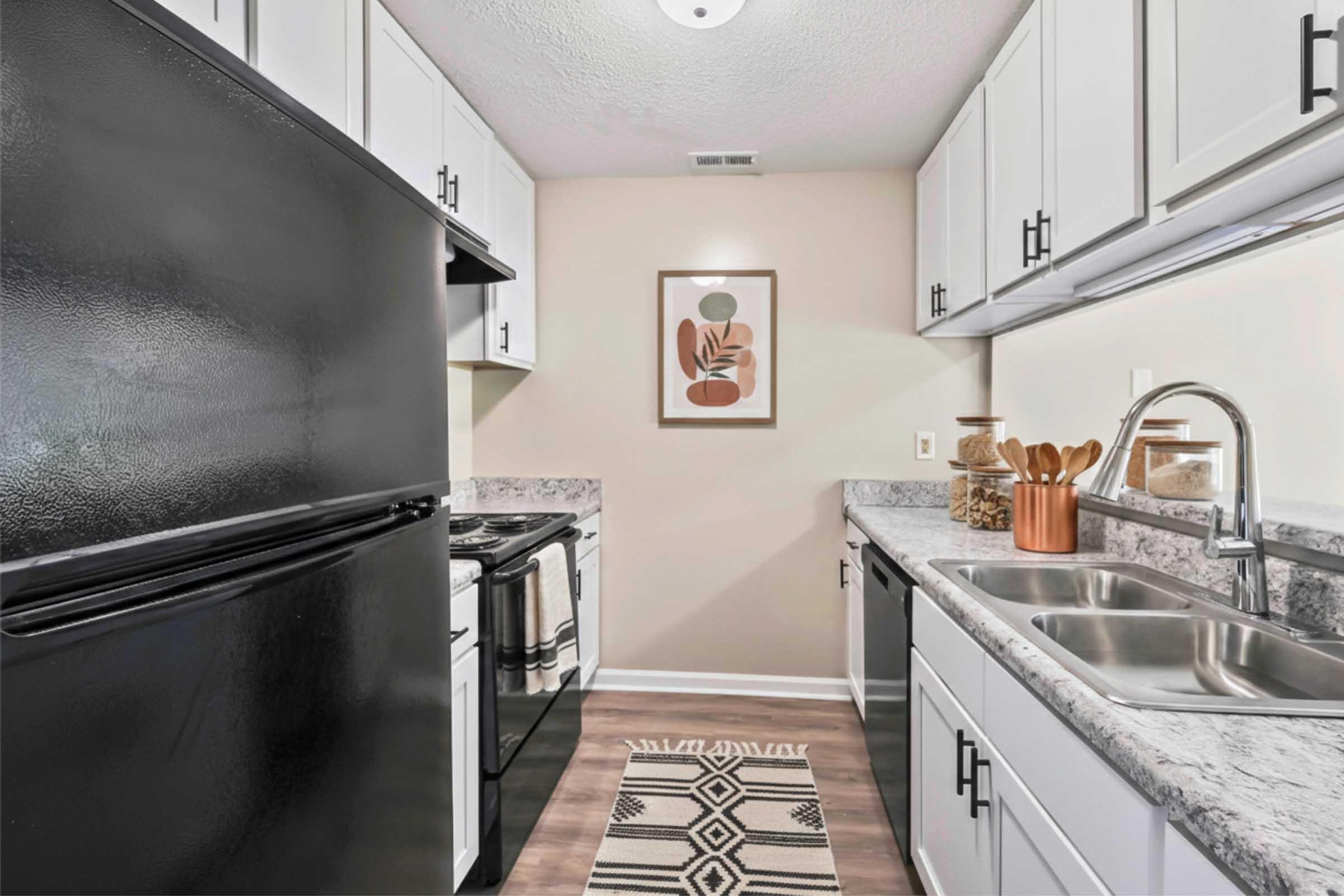 A modern kitchen featuring white cabinets, a black refrigerator, and a black stove. The countertops are made of gray stone, complemented by decorative items and utensils in a wooden holder. A patterned area rug adds warmth, and a framed wall art piece enhances the decor. Bright lighting illuminates the space.