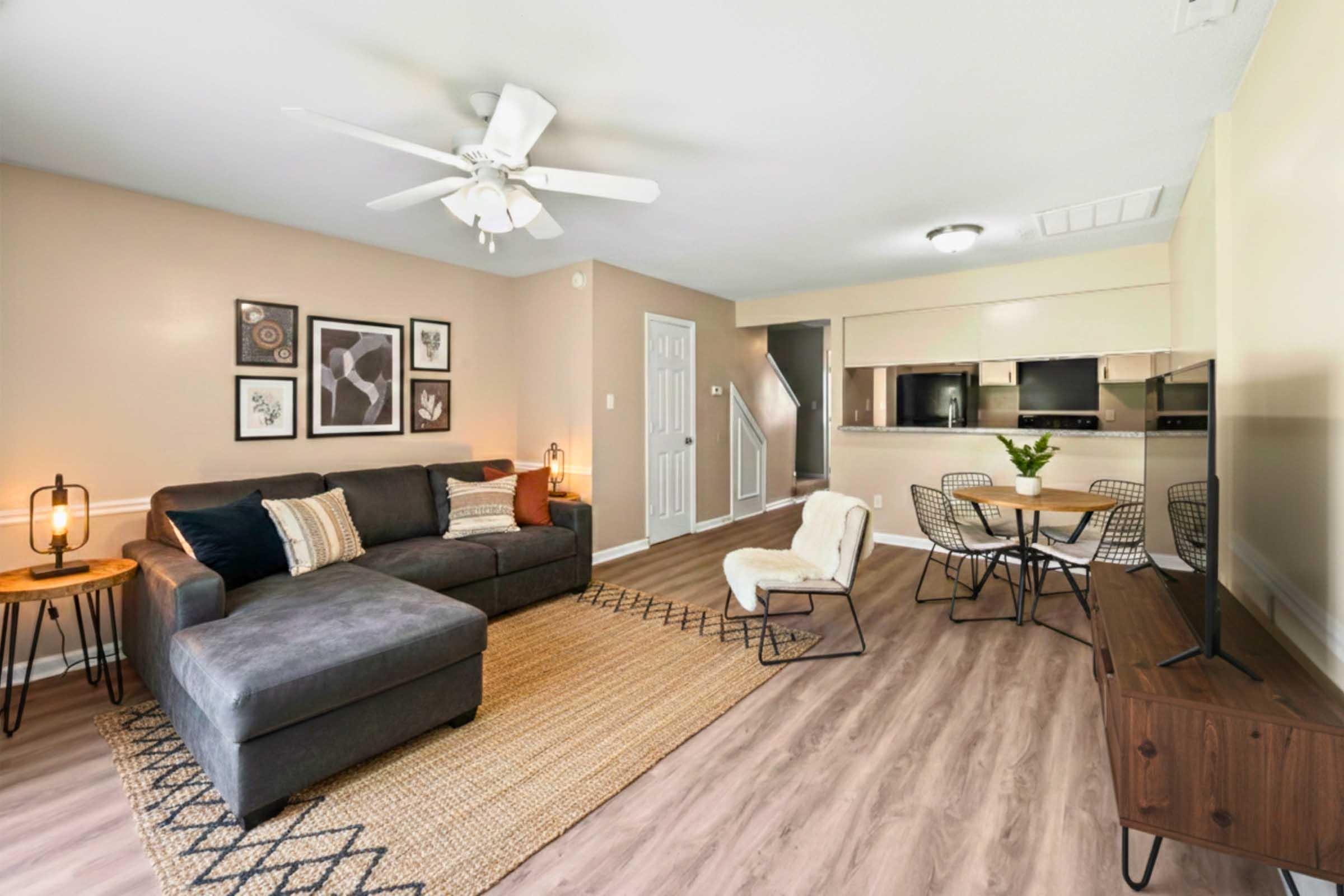 A spacious living room featuring a gray sectional sofa, a decorative rug, and a flat-screen TV. There are decorative wall art pieces, a cozy chair, and a small table with a vase of flowers. The open layout leads to a kitchen area with modern appliances and a staircase in the background. Bright and inviting atmosphere.